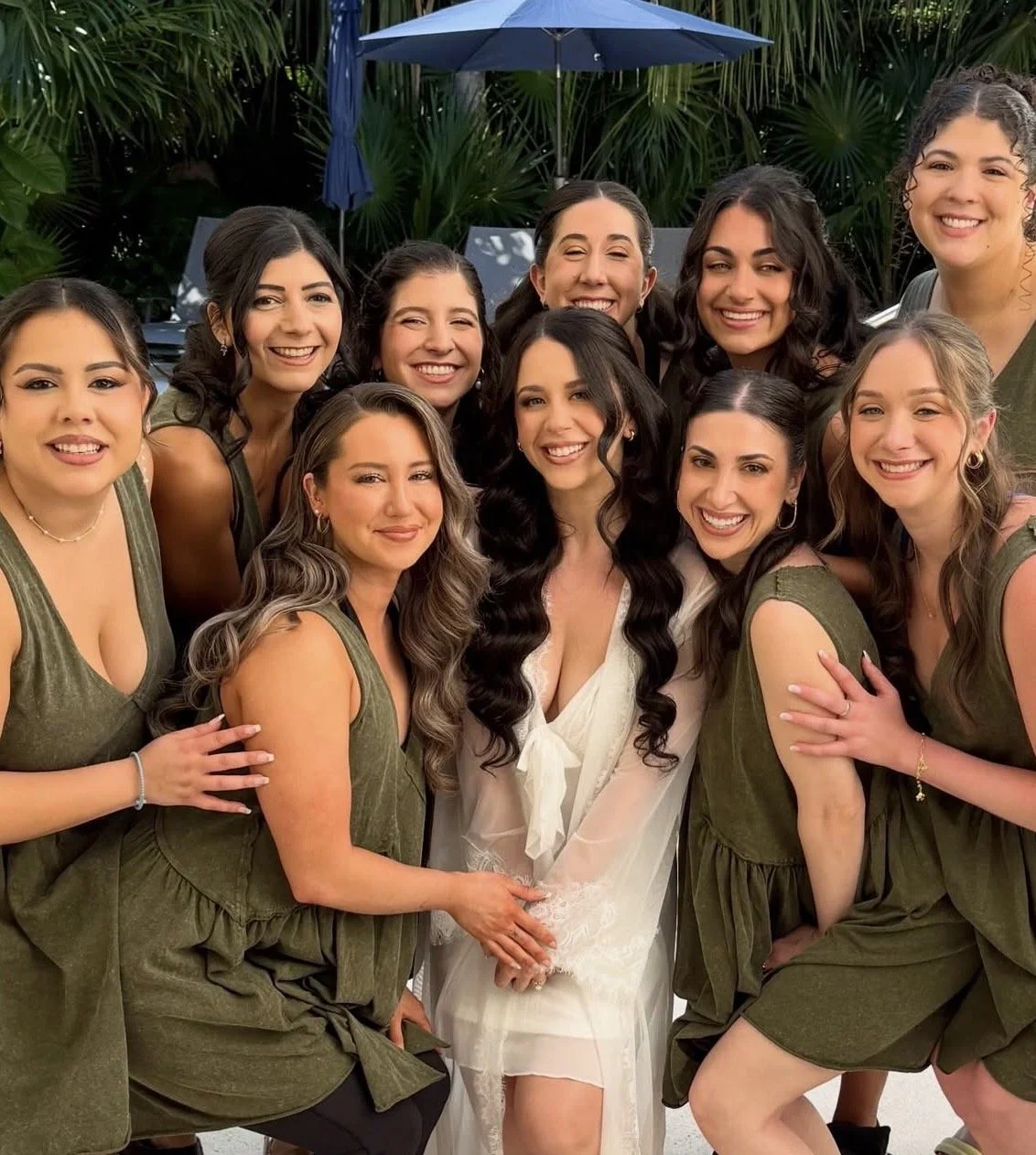 A group of women is smiling and posing together outdoors, with lush green tropical plants and a blue umbrella in the background.
