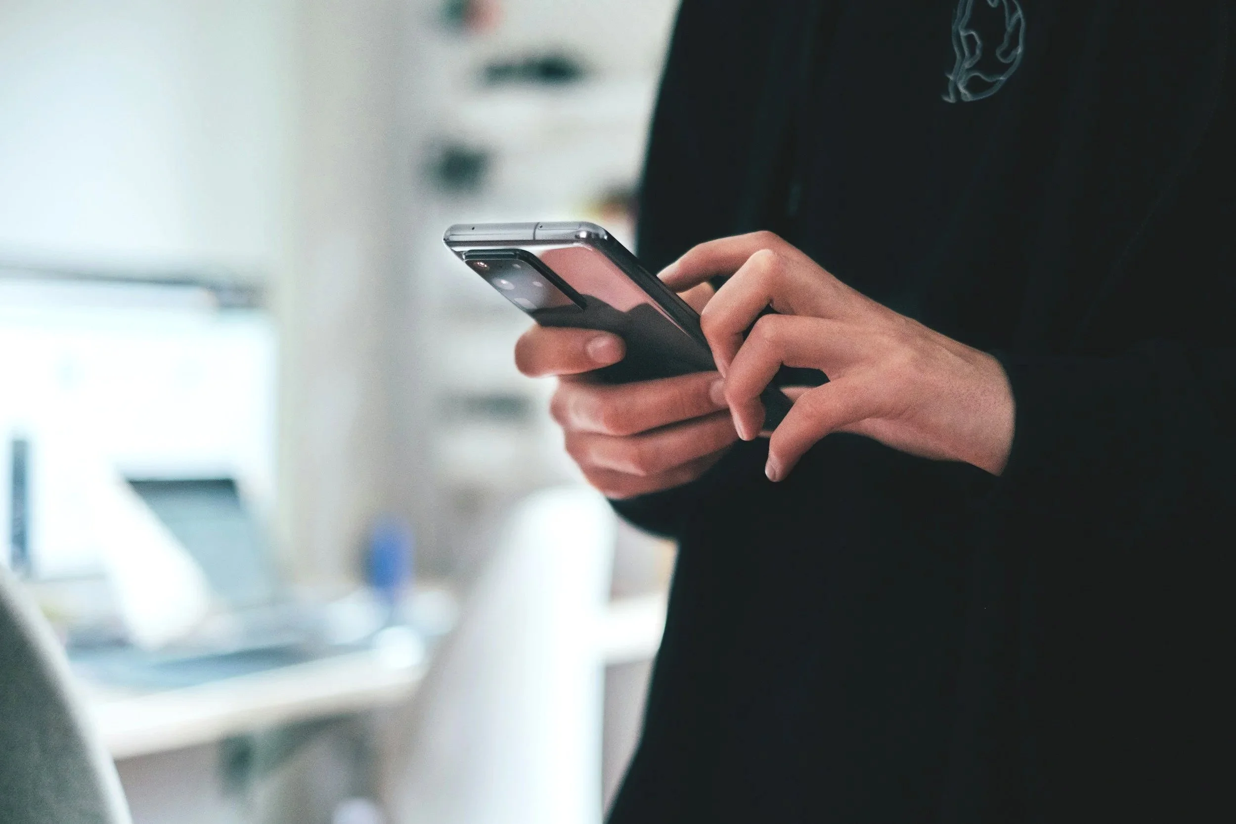 Person holding a smartphone with both hands, wearing a black jacket, in an indoor setting.