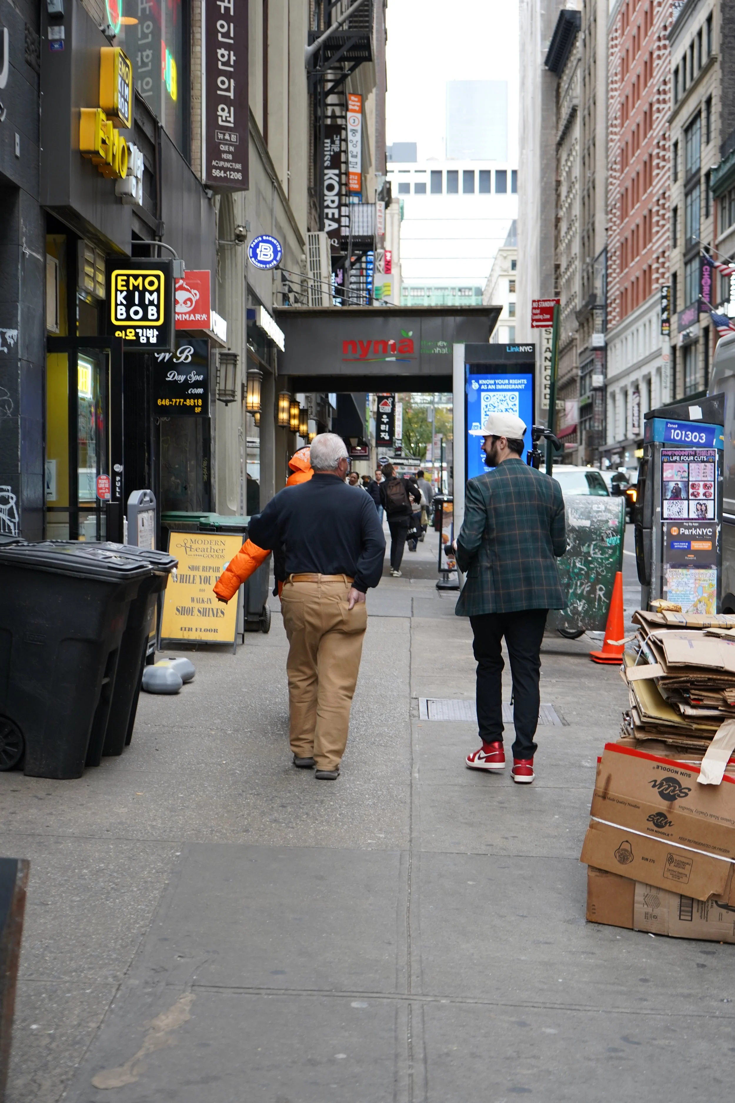 Two men walking on a busy city sidewalk, with various storefronts and signs overhead, including a shoe repair shop and a digital advertising kiosk, and buildings lining the street.