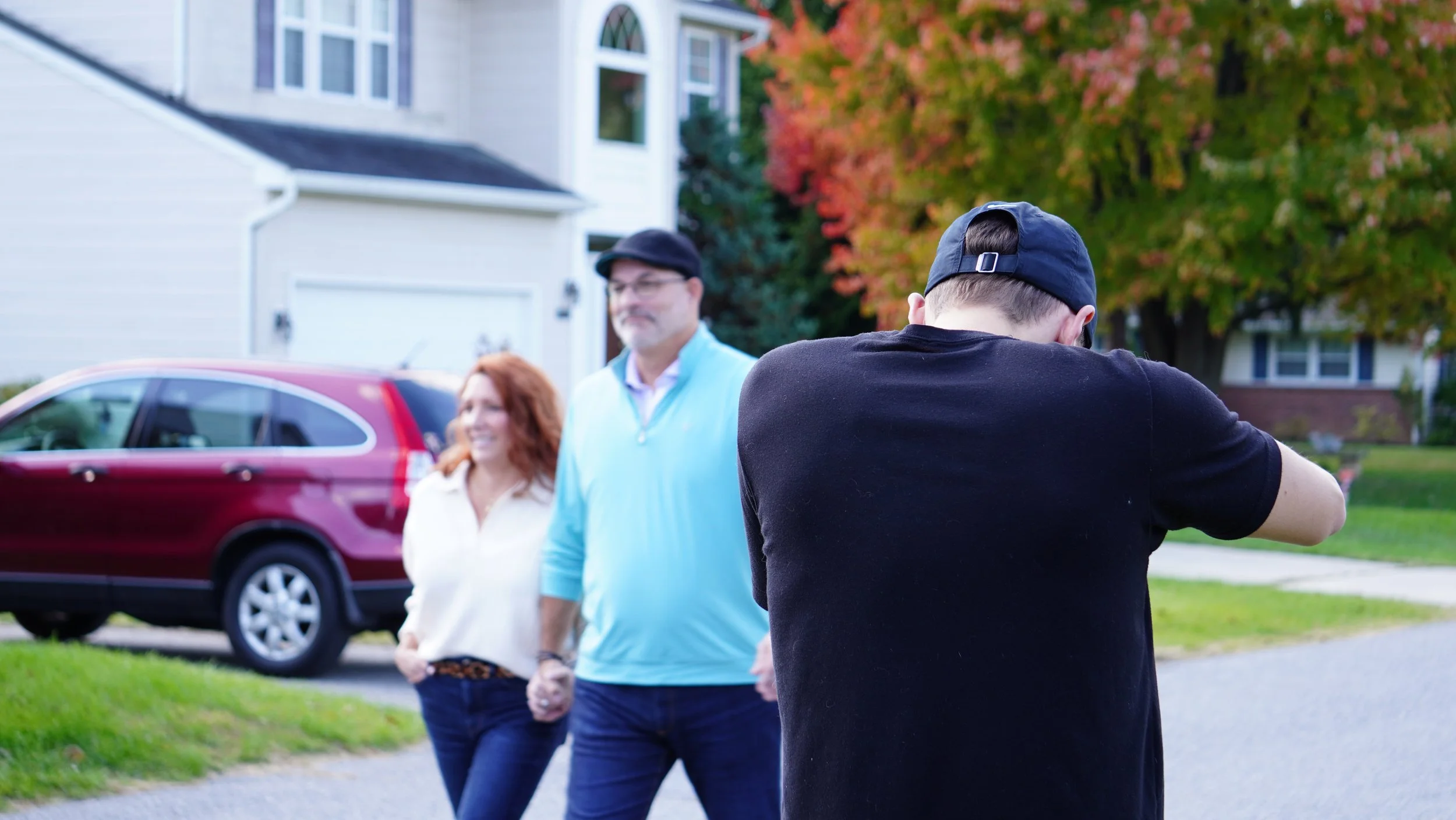 A person with a black shirt and cap taking a photo of a couple holding hands in a suburban neighborhood with trees and houses in the background