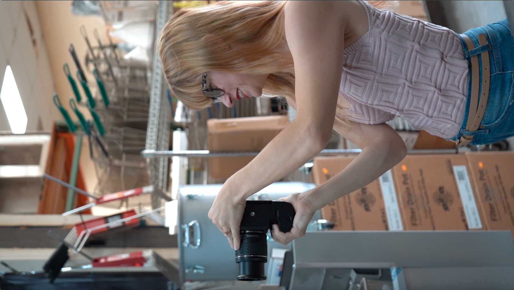 A woman with blonde hair, glasses, and red lipstick, wearing a pink sleeveless top and blue jeans, is operating a camera in a warehouse or storage room filled with boxes and racks.
