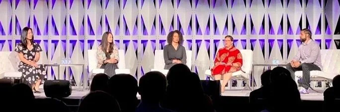 Panel of five women and one man seated on a stage during a discussion or conference, with an audience in the foreground.