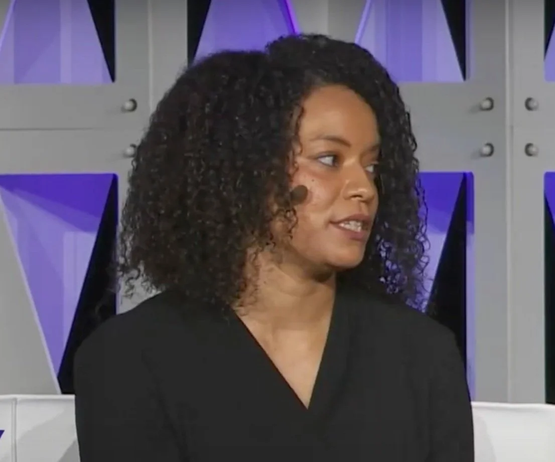 A woman with curly hair talking in a studio setting with modern geometric background decor.