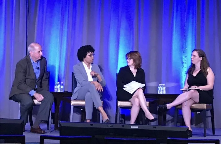 Four people sitting on a stage, engaged in conversation, with blue curtains in the background and bottled water on small tables beside them.