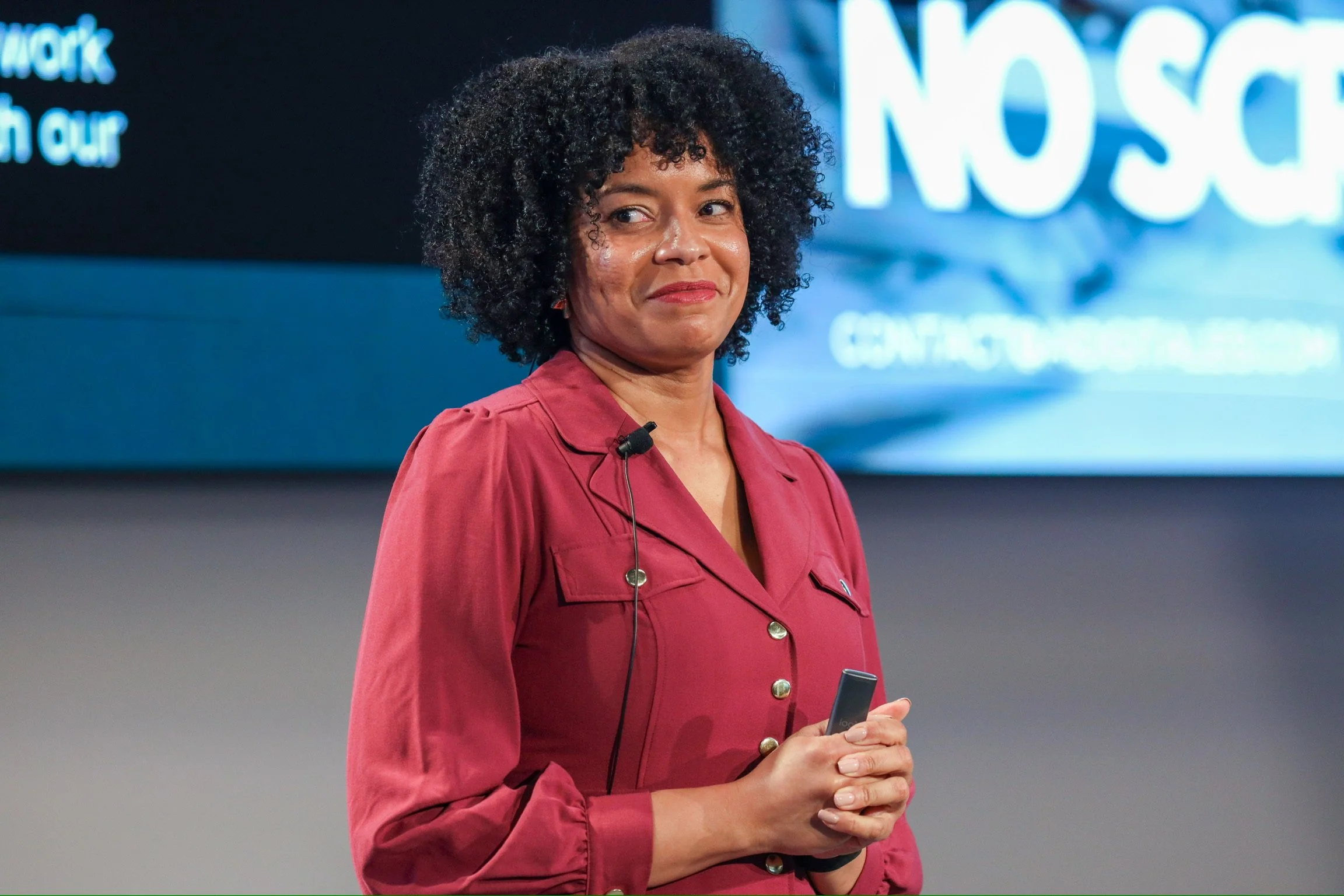 Amber Ivey with curly black hair wearing a fuschia dress holding a microphone in her right hand at business event in California with a large screen behind her.