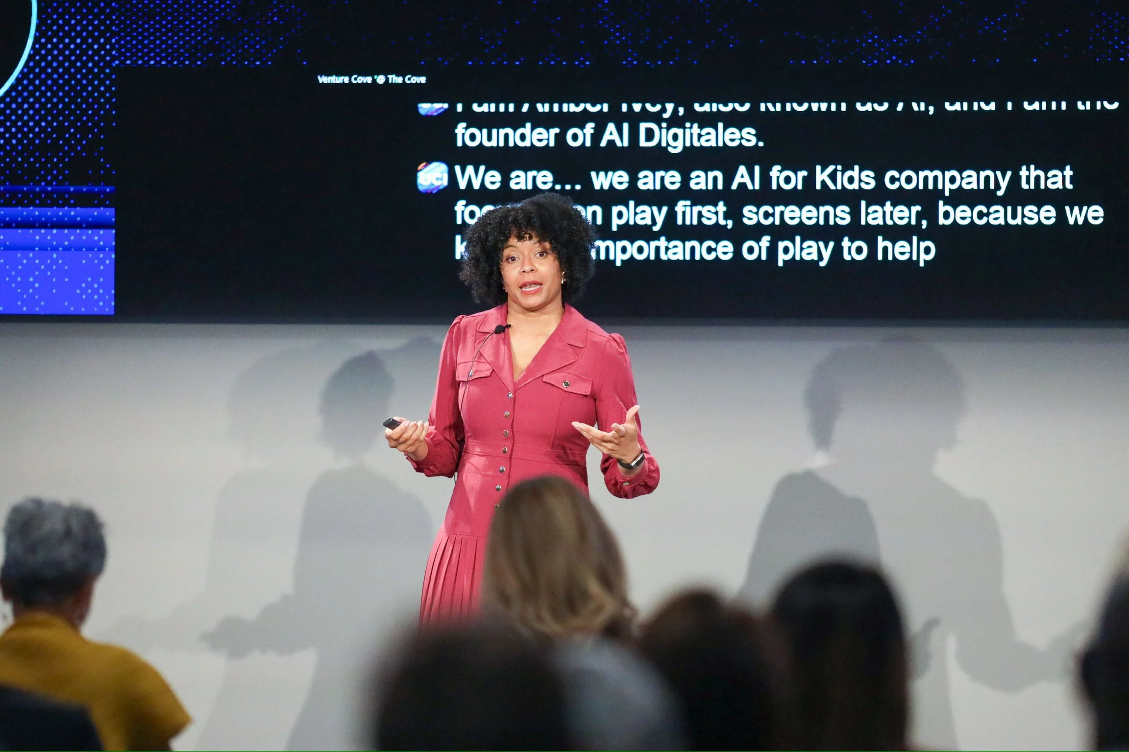 A woman with curly black hair wearing a red dress speaking in front of a large screen during a presentation, with an audience in the foreground.