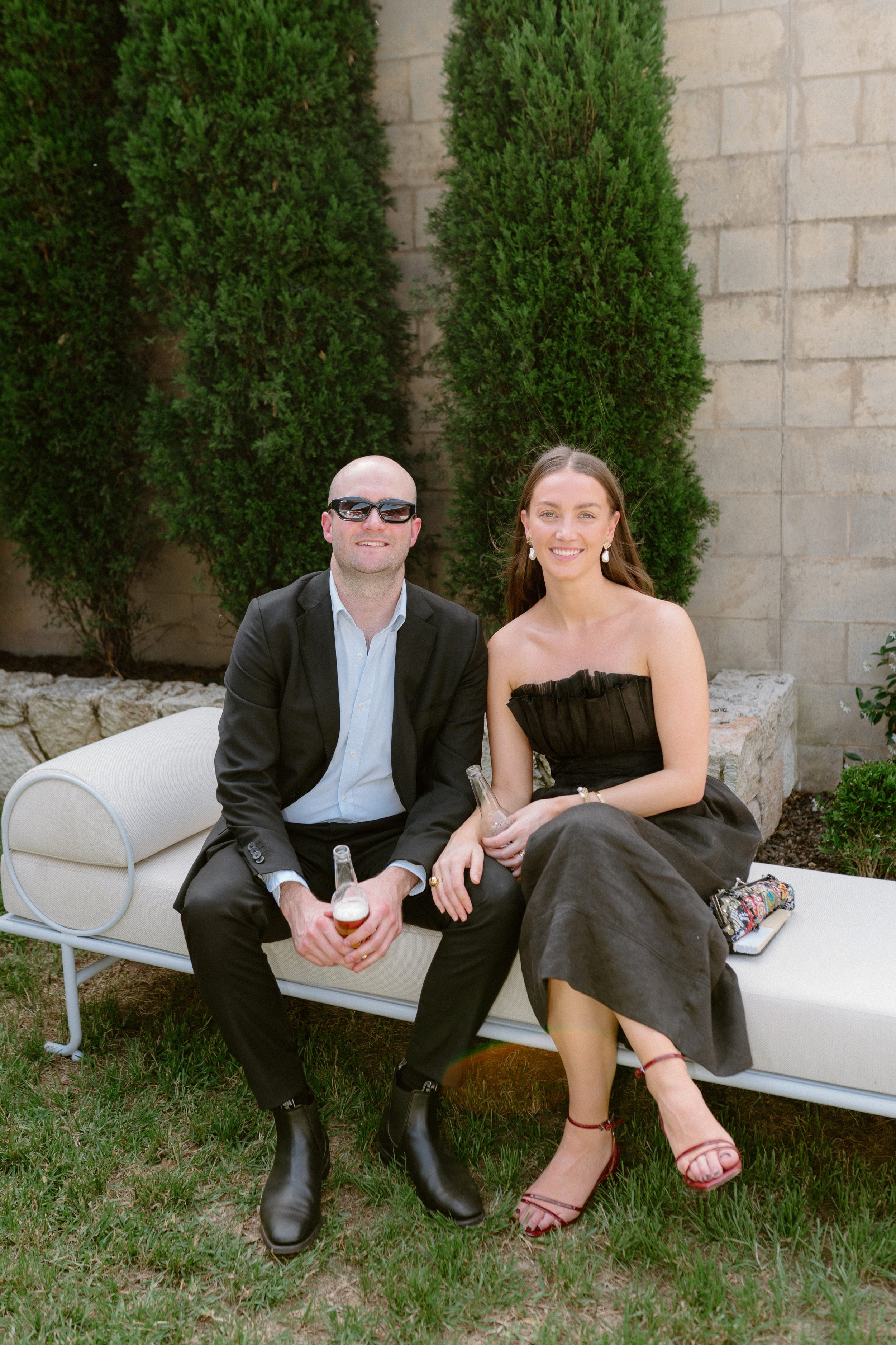 A man and a woman sitting on a white bench outdoors against a stone wall with tall greenery. The man is wearing a black suit, white shirt, black boots, and sunglasses. The woman is wearing a strapless black dress, red high heels, and is holding a gla