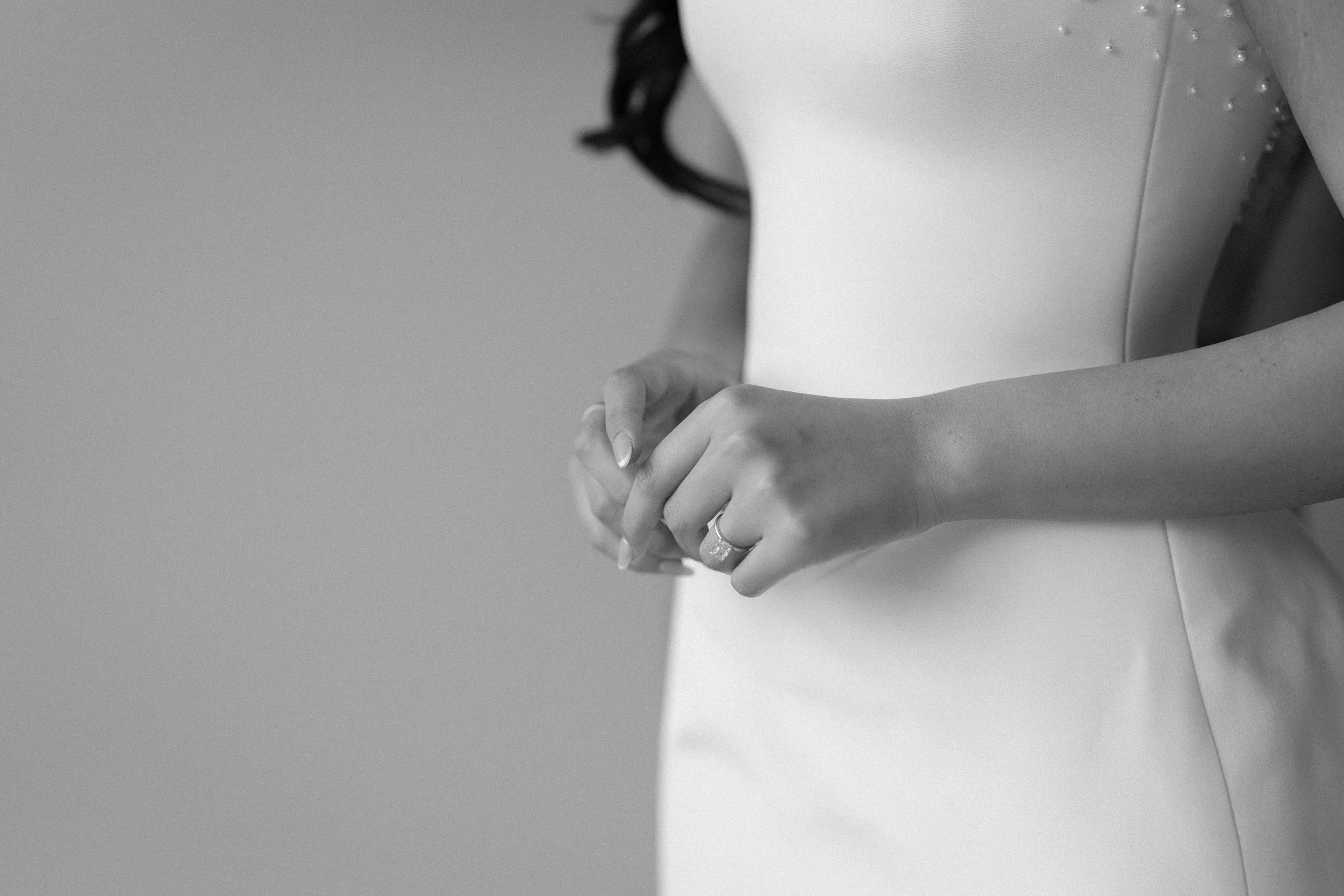 Close-up of a woman in a white dress with pearl embellishments, holding her hands together with a wedding ring on her finger, in black and white.