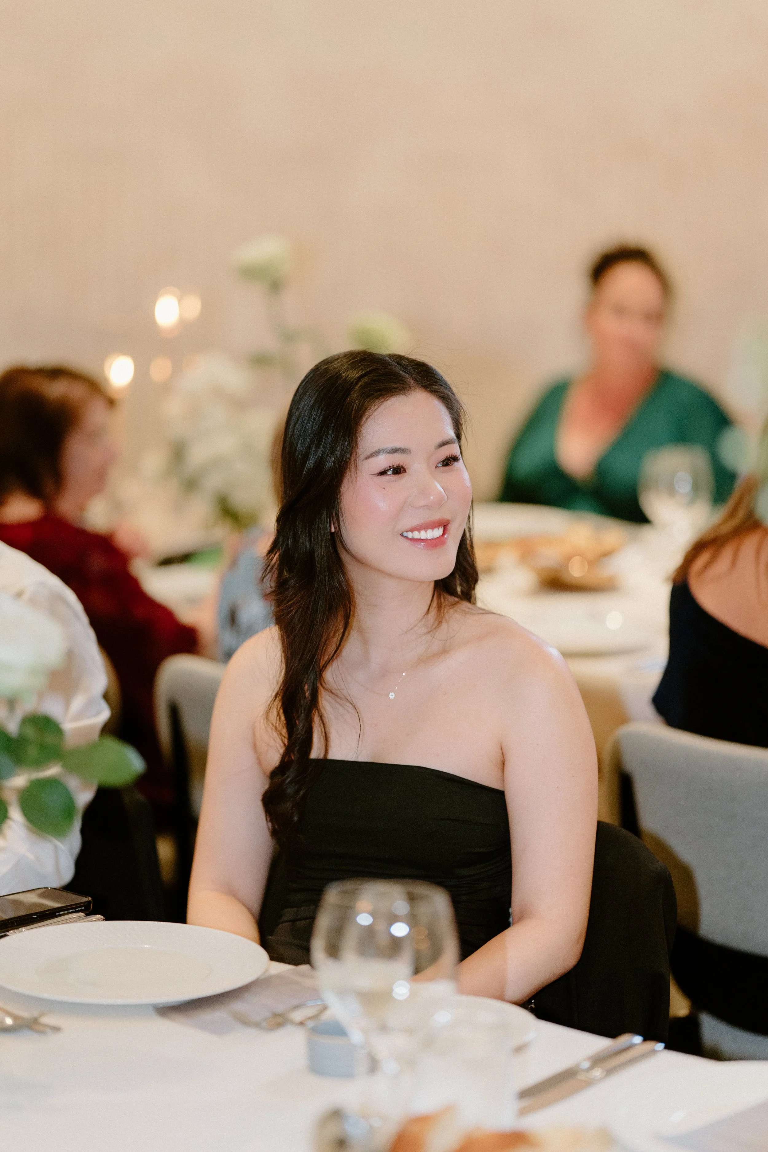 A woman with dark hair in loose waves, wearing a black strapless dress, smiling at a formal event with other guests seated at tables with white tablecloths, glassware, and flower arrangements.