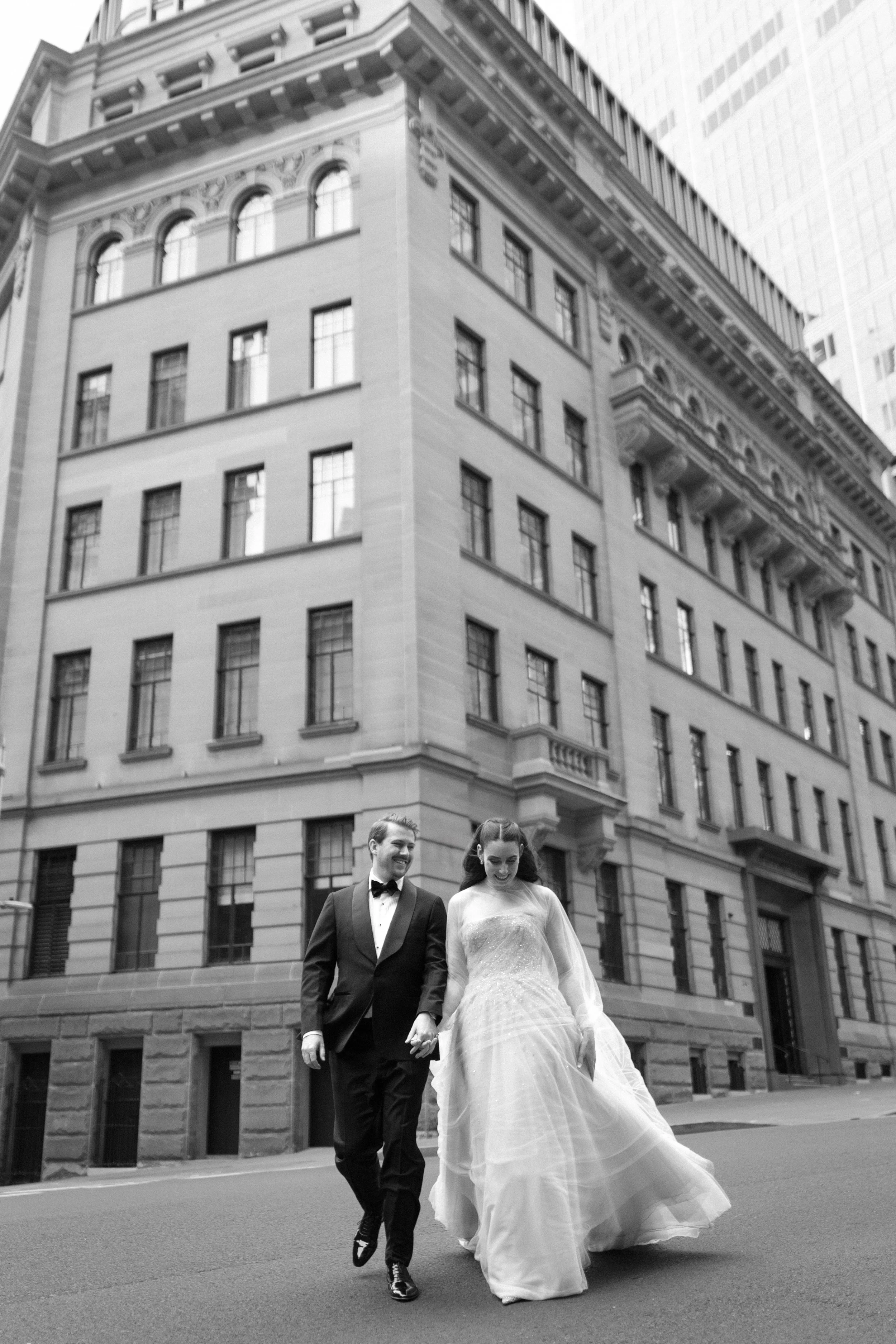 A happy couple, dressed in wedding attire, walking hand in hand on a city street with tall buildings in the background.