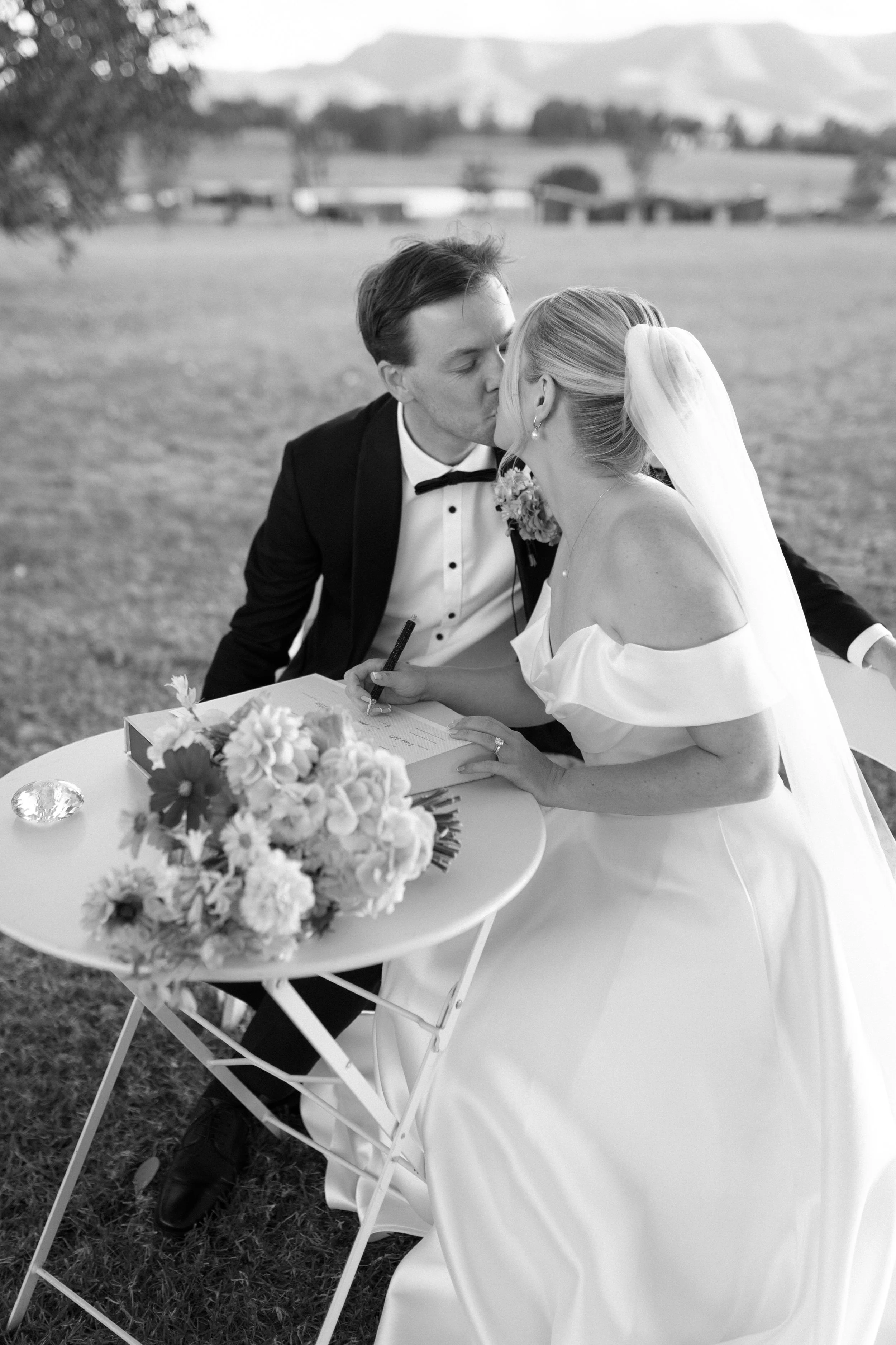 A black-and-white photo of a bride and groom sharing a kiss outdoors at a wedding ceremony. The bride is wearing a white off-the-shoulder wedding dress and a veil, while the groom is dressed in a tuxedo. There is a table with a bouquet of flowers in 