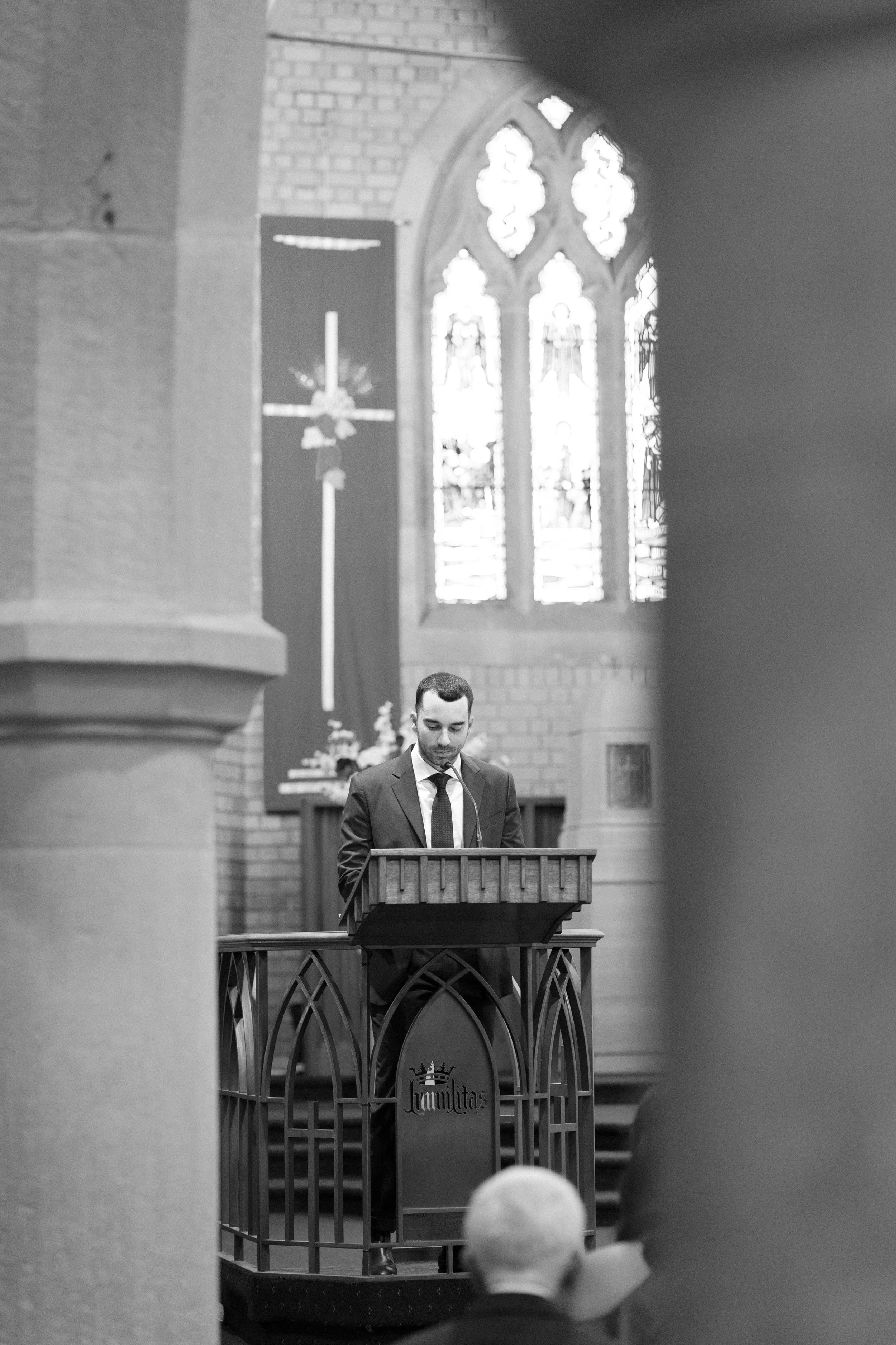 A man in a suit standing at a pulpit inside a church, with stained glass windows and Gothic architecture visible in the background.