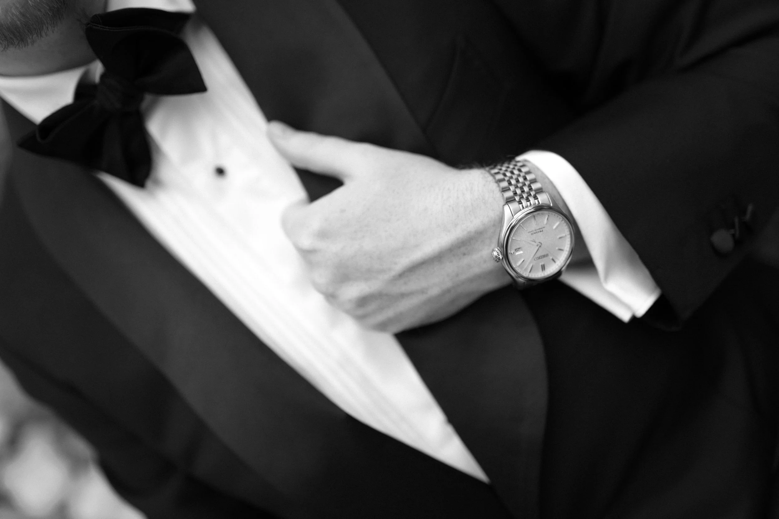 Close-up of a man adjusting his tuxedo shirt cuff, wearing a wristwatch, dressed formally in a tuxedo with a bow tie.