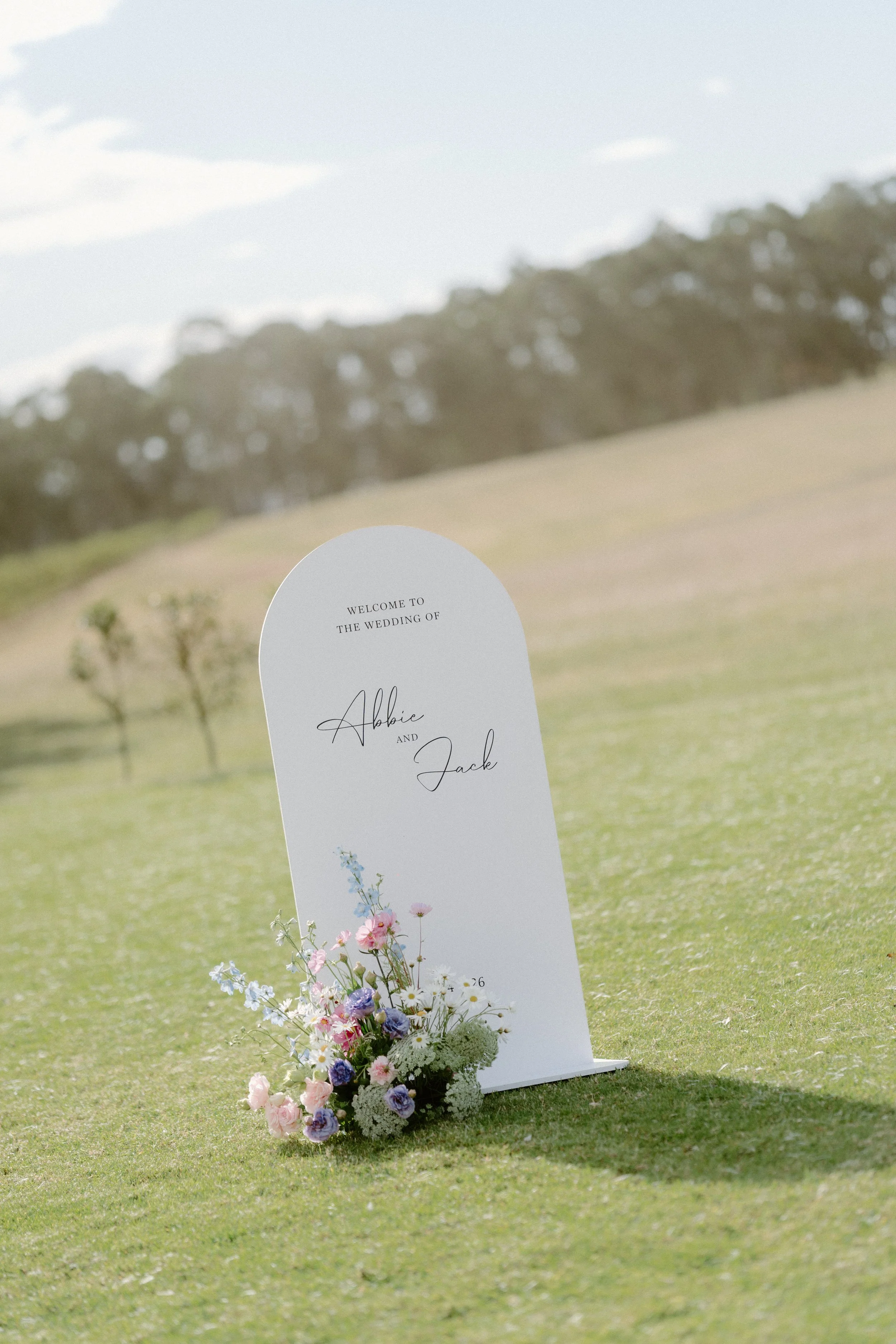 Outdoor wedding sign with floral arrangements on green grass with trees and cloudy sky in background.