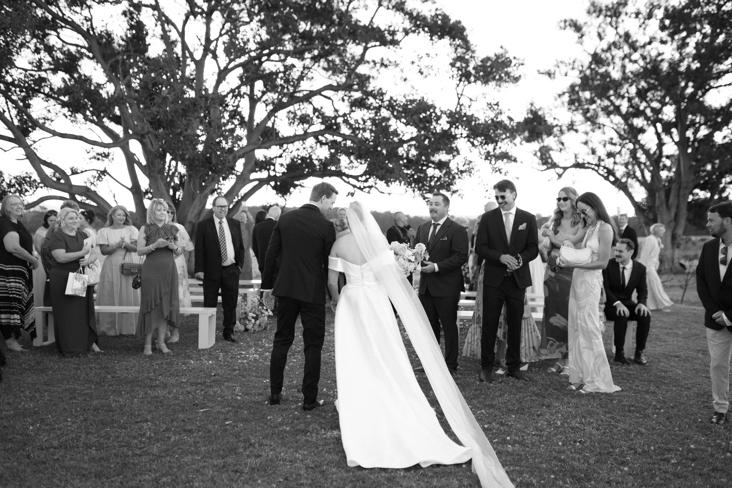 A wedding ceremony outdoors under a large tree, with the bride and groom in the center exchanging vows surrounded by guests. Bimbadgen Palmers Lane Wedding, Hunter Valley