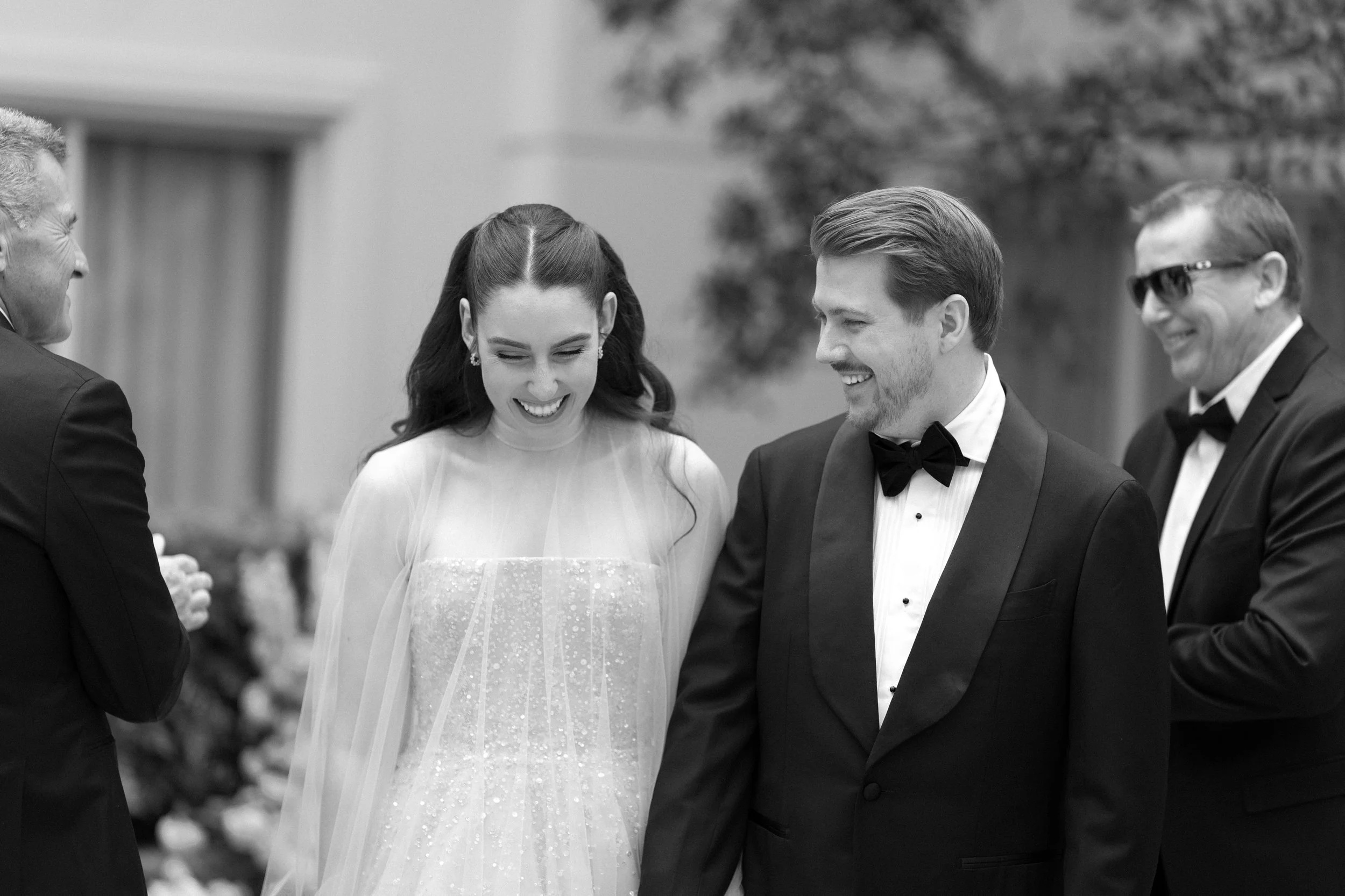 Black and white photo of a wedding scene with smiling bride and groom dressed in wedding attire, surrounded by groomsmen.