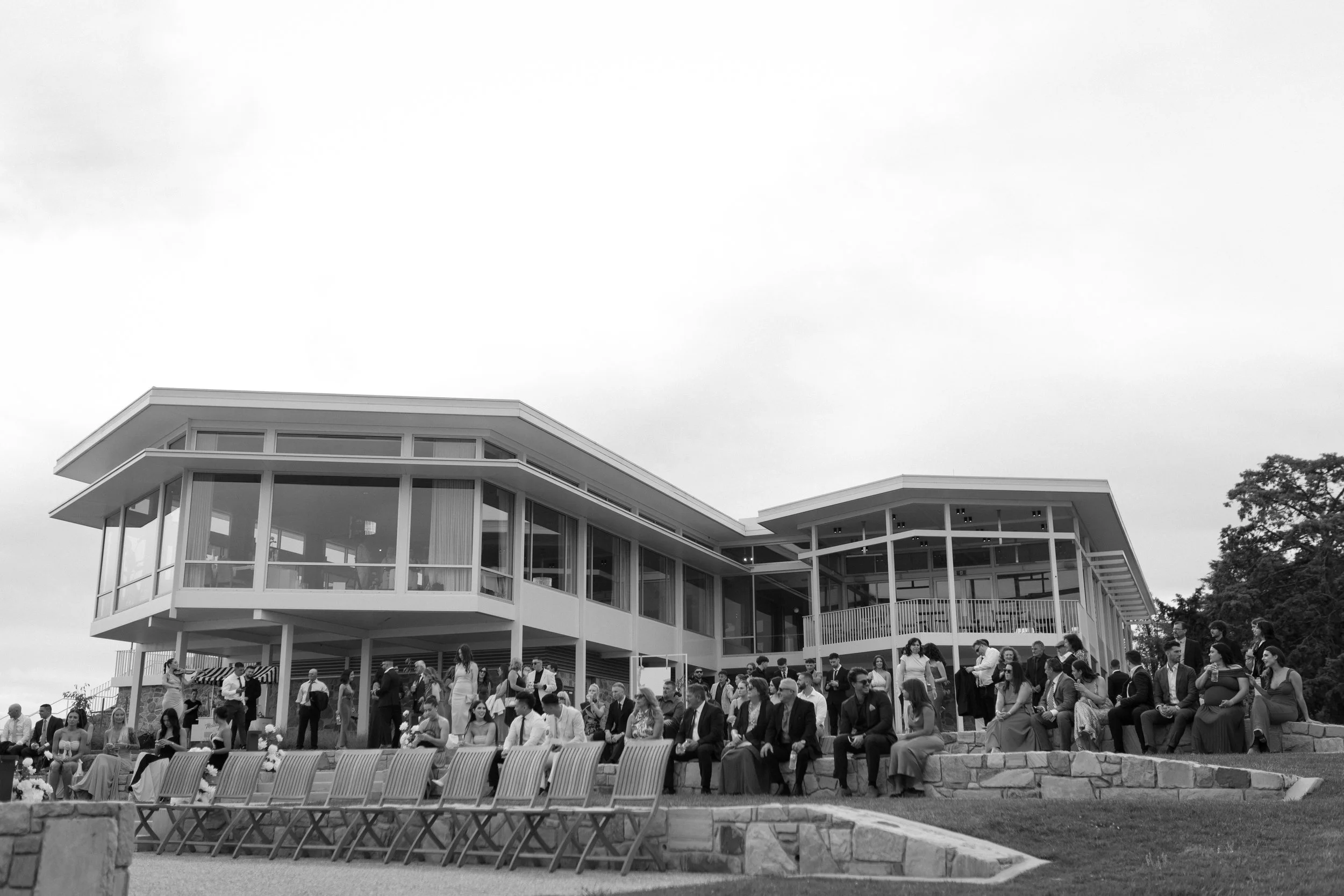 Guests seated outside a modern building with large glass windows, attending a formal event or wedding.