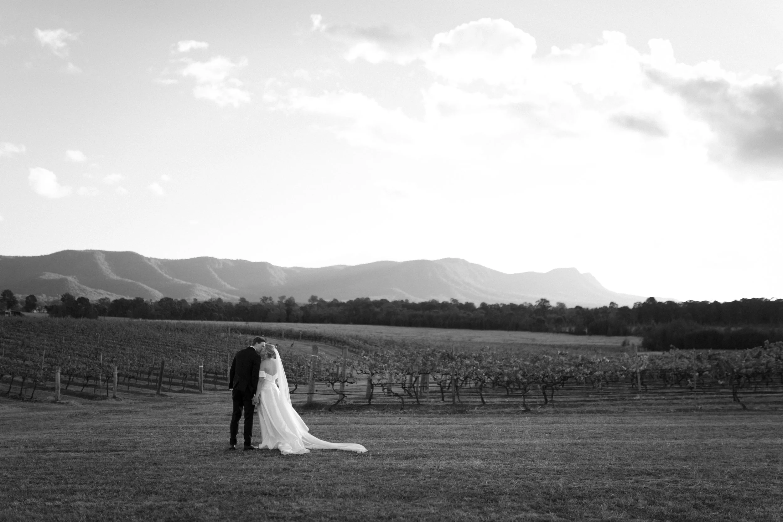 A black and white photo of a bride and groom standing on a grassy field in front of a vineyard with mountains in the background. The groom is kissing the bride on the forehead while they hold hands. Bimbadgen Palmers Lane Wedding, Hunter Valley