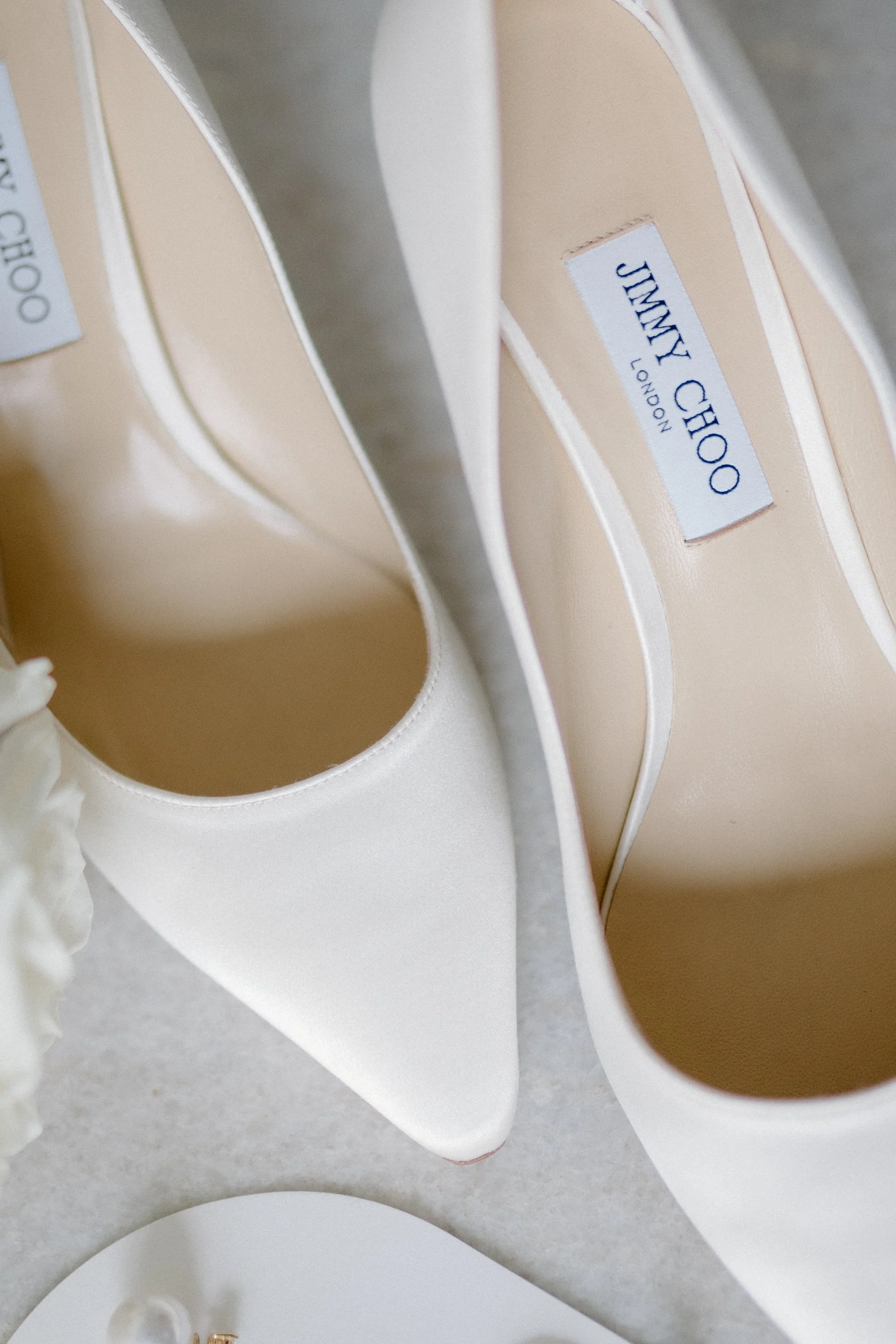Close-up of white Jimmy Choo high-heeled shoes with pointed toes, placed on a light surface.