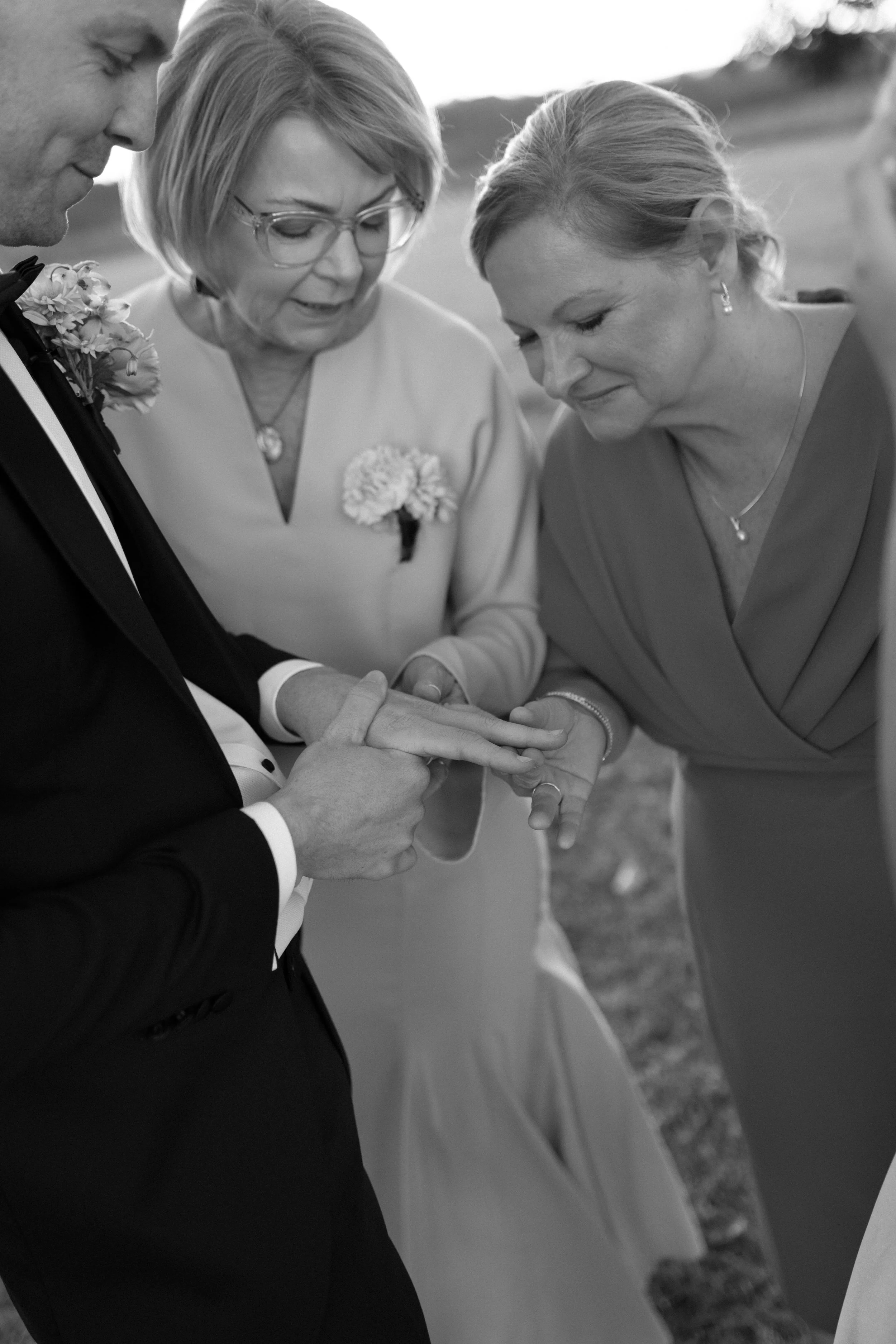 A black and white photo of a wedding ceremony showing a man in a suit and a woman in a dress holding hands, with a woman in glasses and a boutonniere officiating the ceremony.