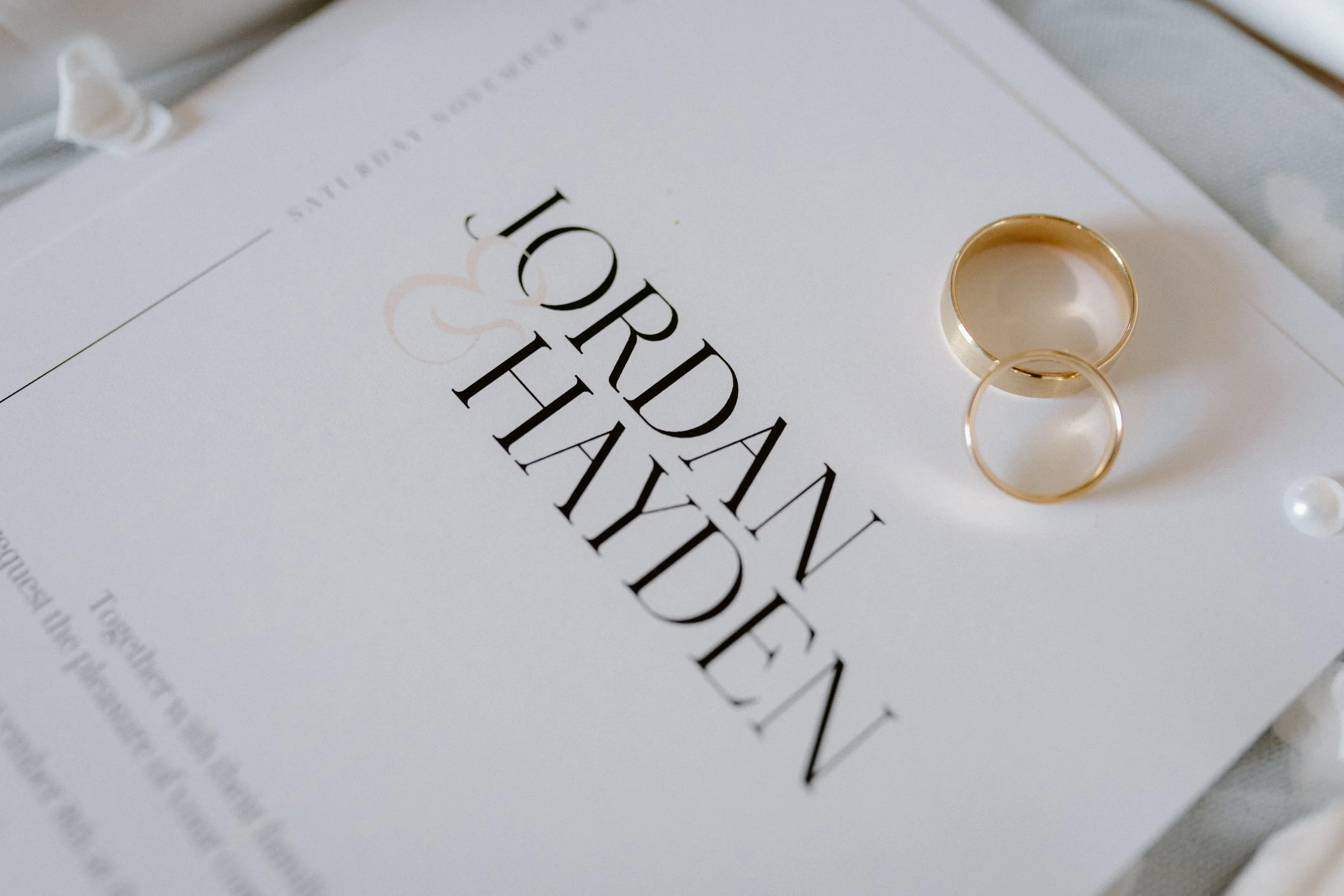 A wedding invitation with the words "Wedding Invitation" and a fleur-de-lis. Gold rings are placed on top of the invitation, and there are pearl and white fabric decorations nearby.