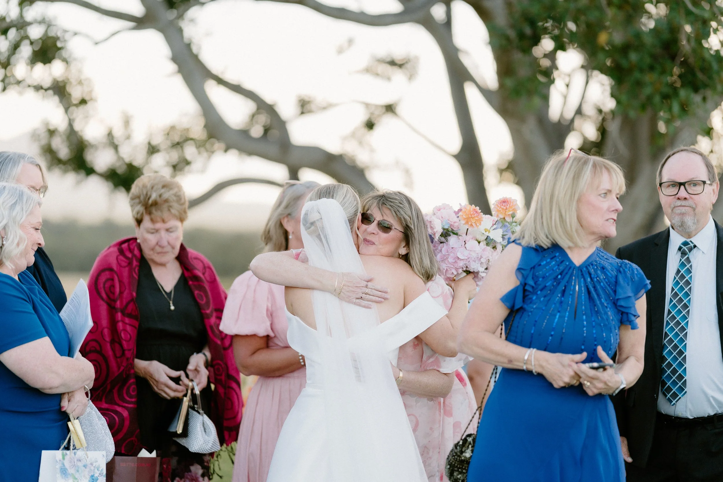A group of people, including women and men, gathered outdoors for a wedding ceremony, with a woman in a white dress hugging another woman, possibly the bride, amidst trees and bright sunlight.