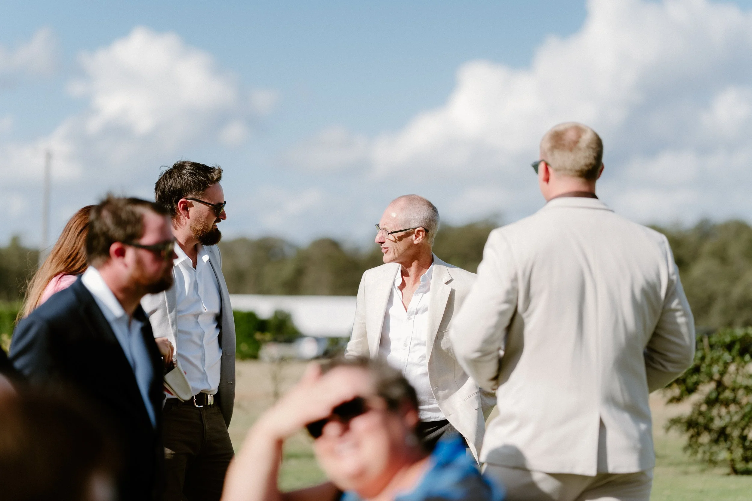 Group of people gathered outdoors, engaged in conversation, with a backdrop of blue sky and clouds.