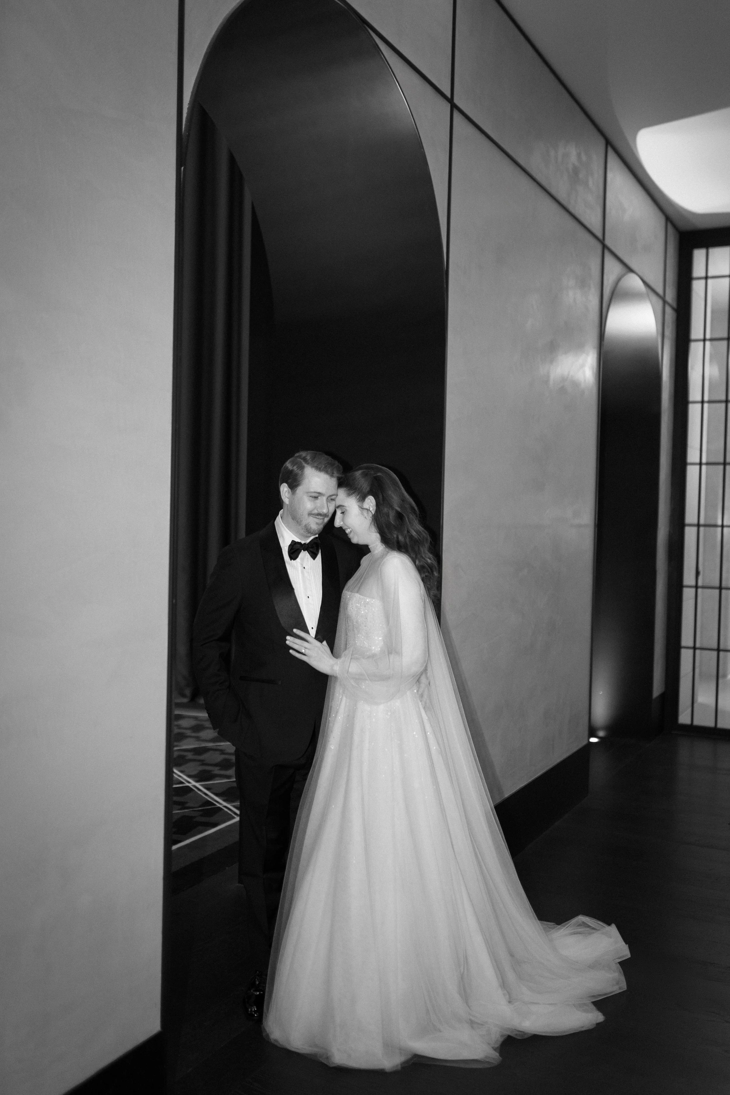 A black-and-white photo of a bride and groom smiling and leaning their foreheads together in a modern indoor setting.