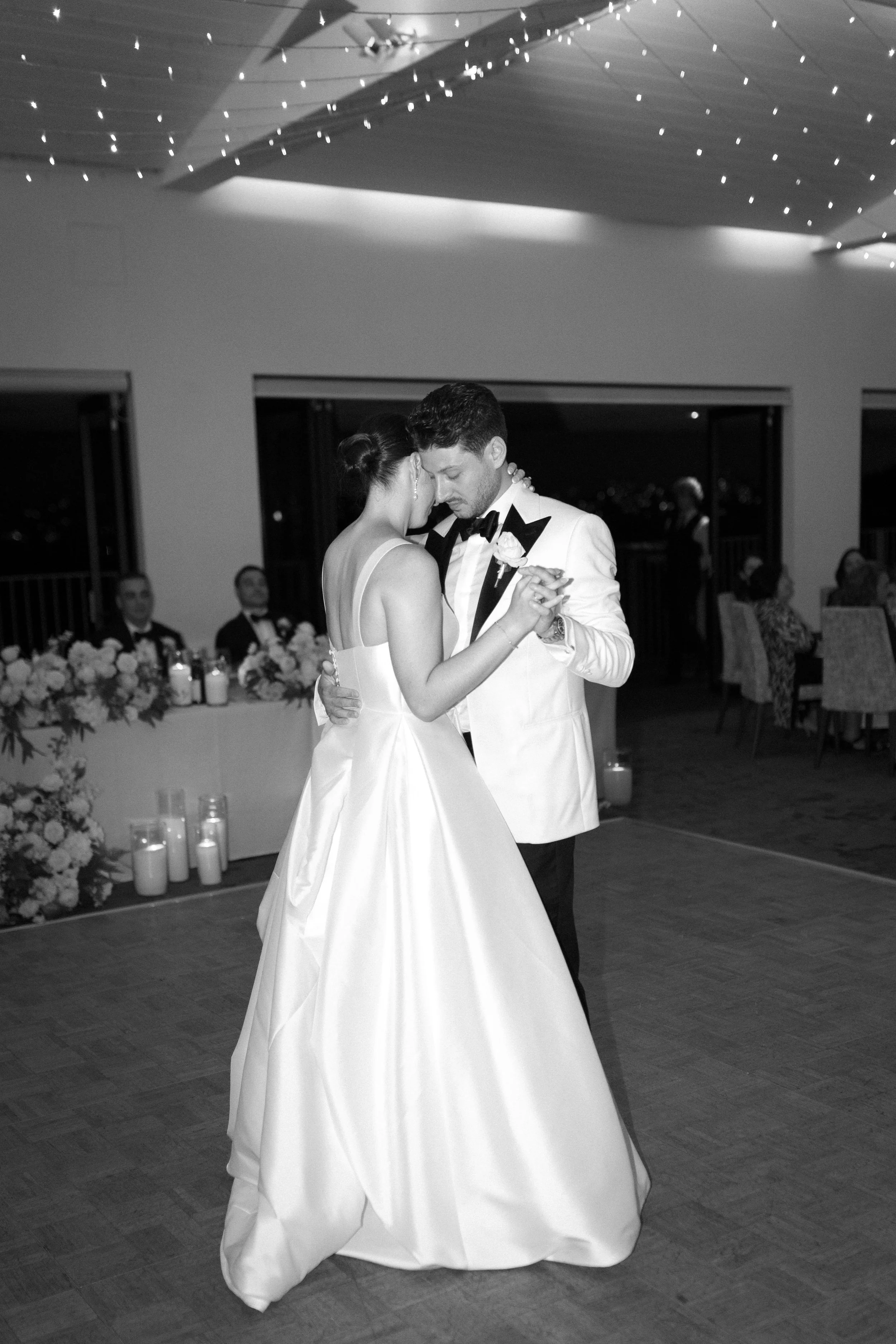 Sergeants Mess, Mosman Wedding Venue. Sergeants Mess Wedding Photos. A black and white photo of a bride and groom sharing their first dance at a wedding reception, with guests seated at tables in the background.