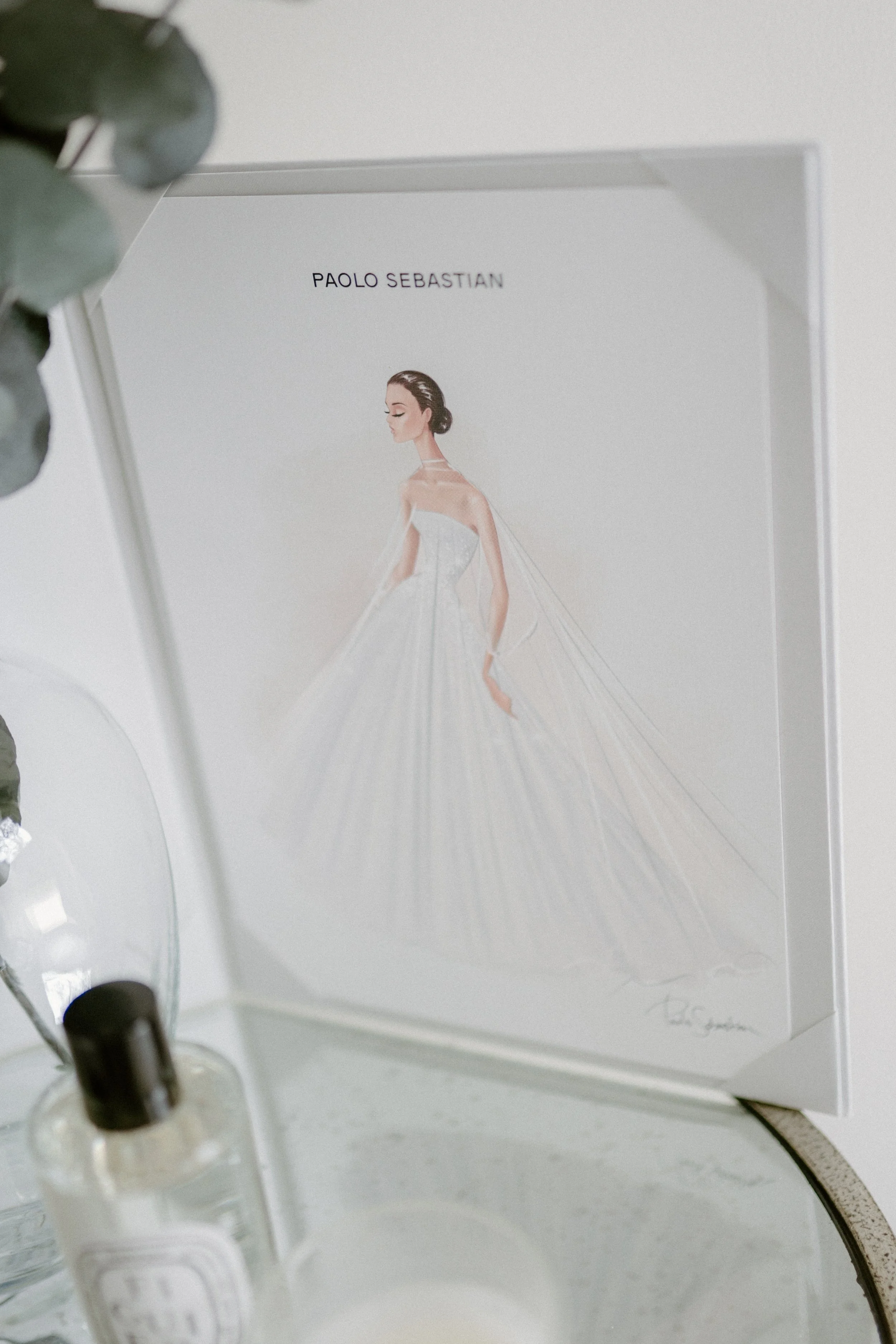 A framed fashion illustration featuring a woman in a strapless white gown with a full skirt, displayed on a tabletop next to a small bottle and a glass dish with a plant branch.