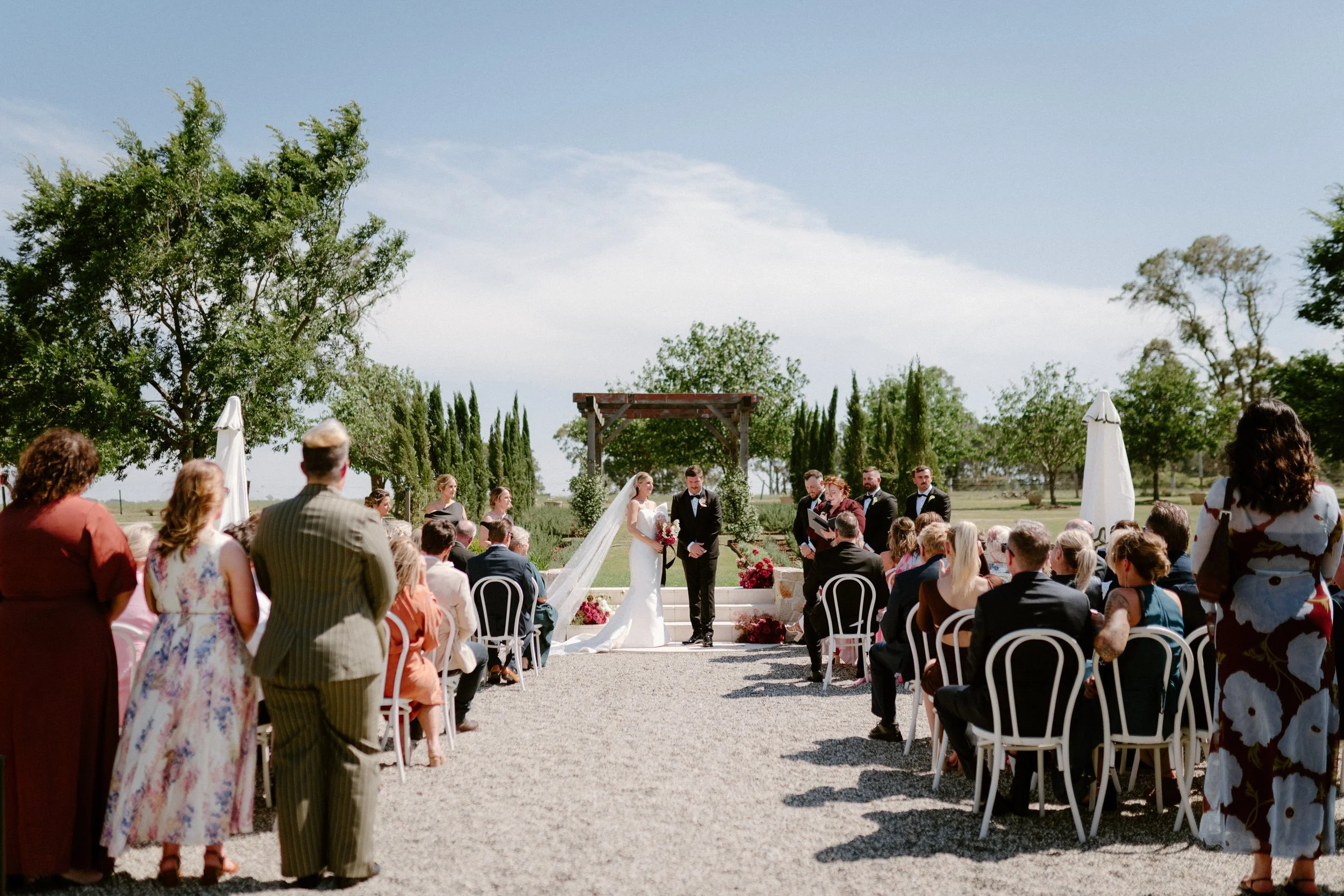 Outdoor wedding ceremony with bride and groom at altar, seated guests, officiant, on a sunny day with trees in the background.