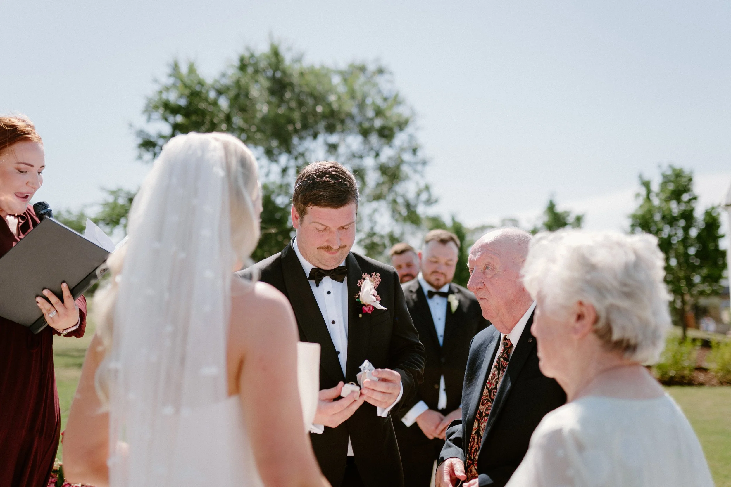 Wedding ceremony outdoors with the bride, groom, and elderly couple exchanging vows, officiant on the left reading from a book, guests in the background, sunny day with trees.