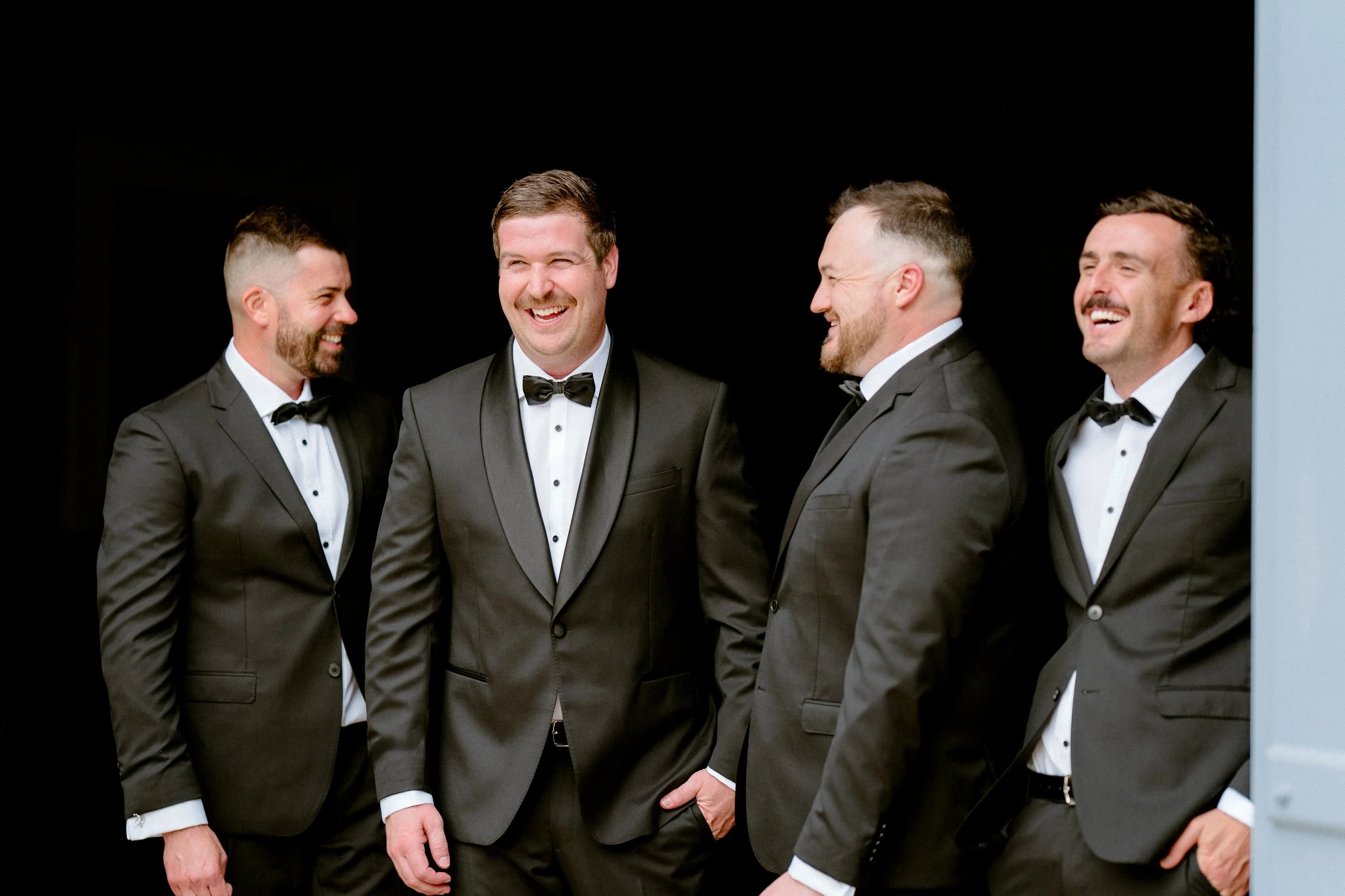 Five men in tuxedos laughing and smiling together.