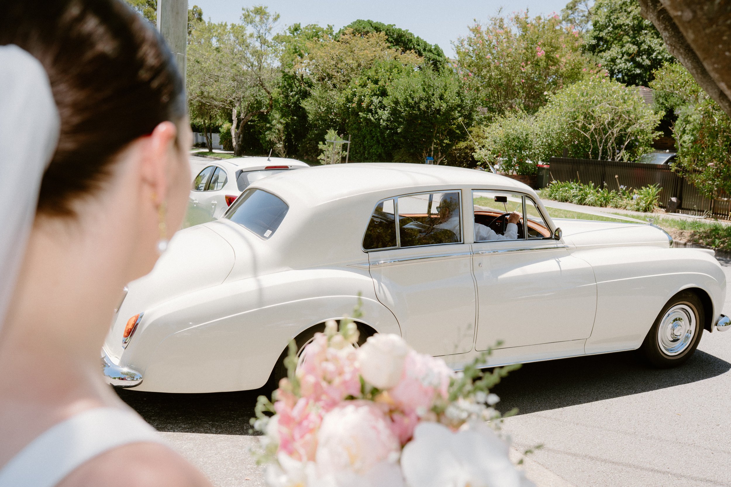 A bride holding a bouquet of pink and white flowers looks at a vintage white car driving on a suburban street with green trees and parked cars.