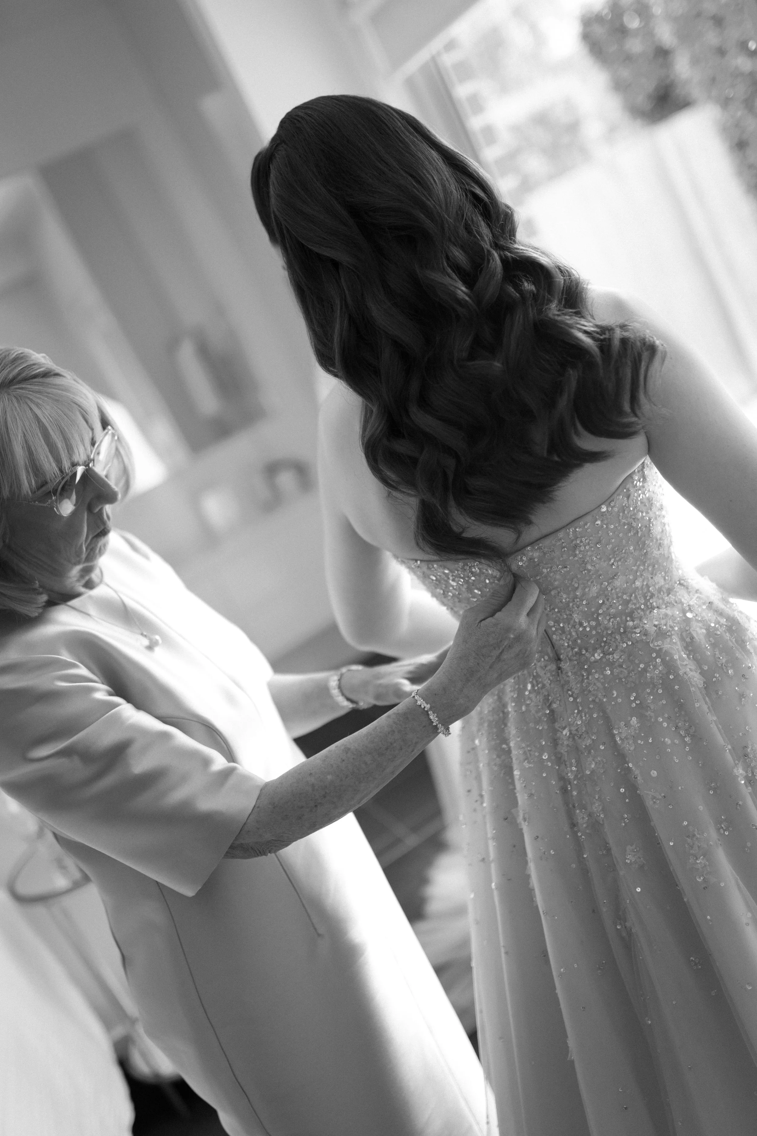 An elderly woman helping a bride with her wedding dress in a room with a large window.