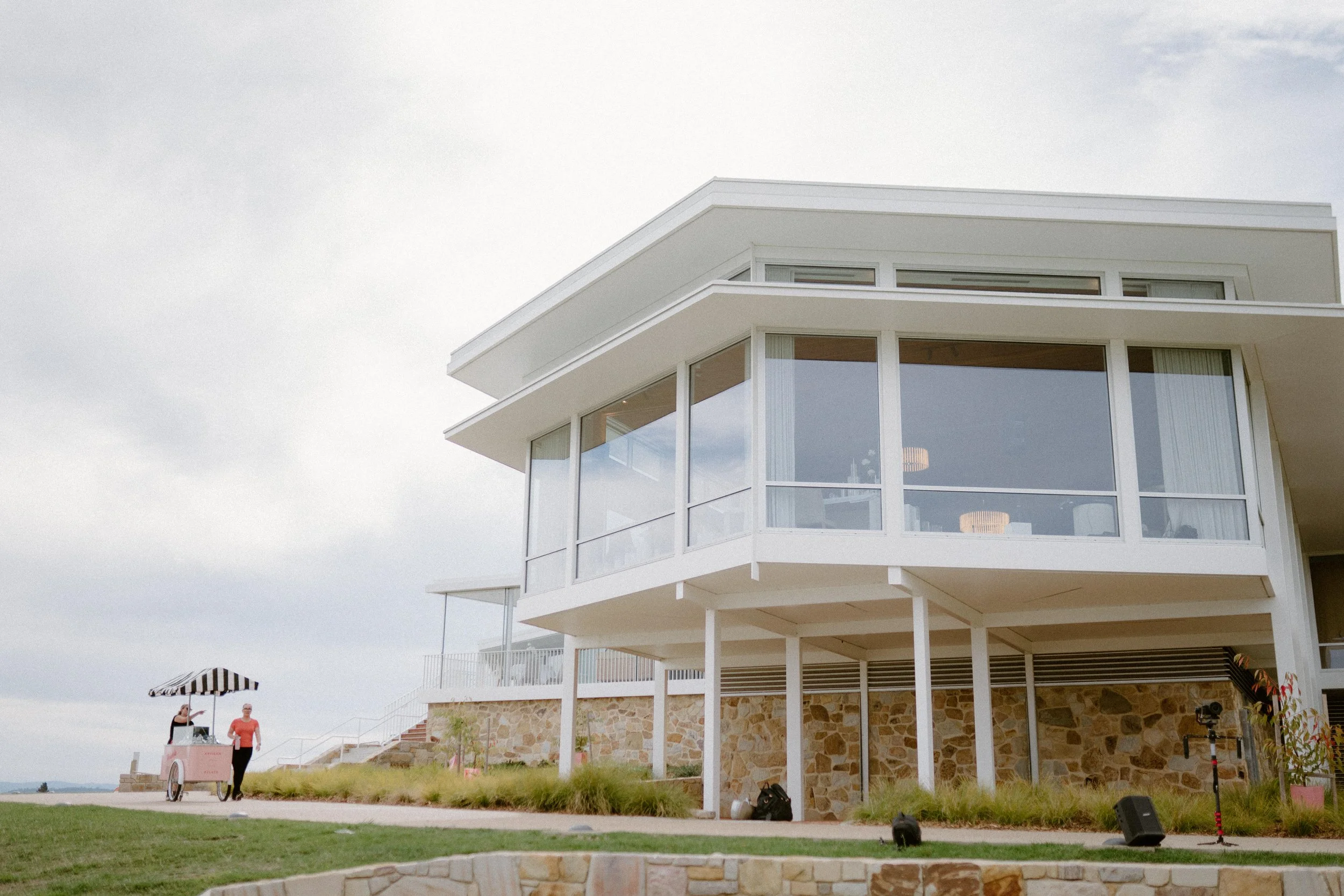 Modern multi-story house with large glass windows and a stone foundation, situated outdoors with a grassy area and a cloudy sky.