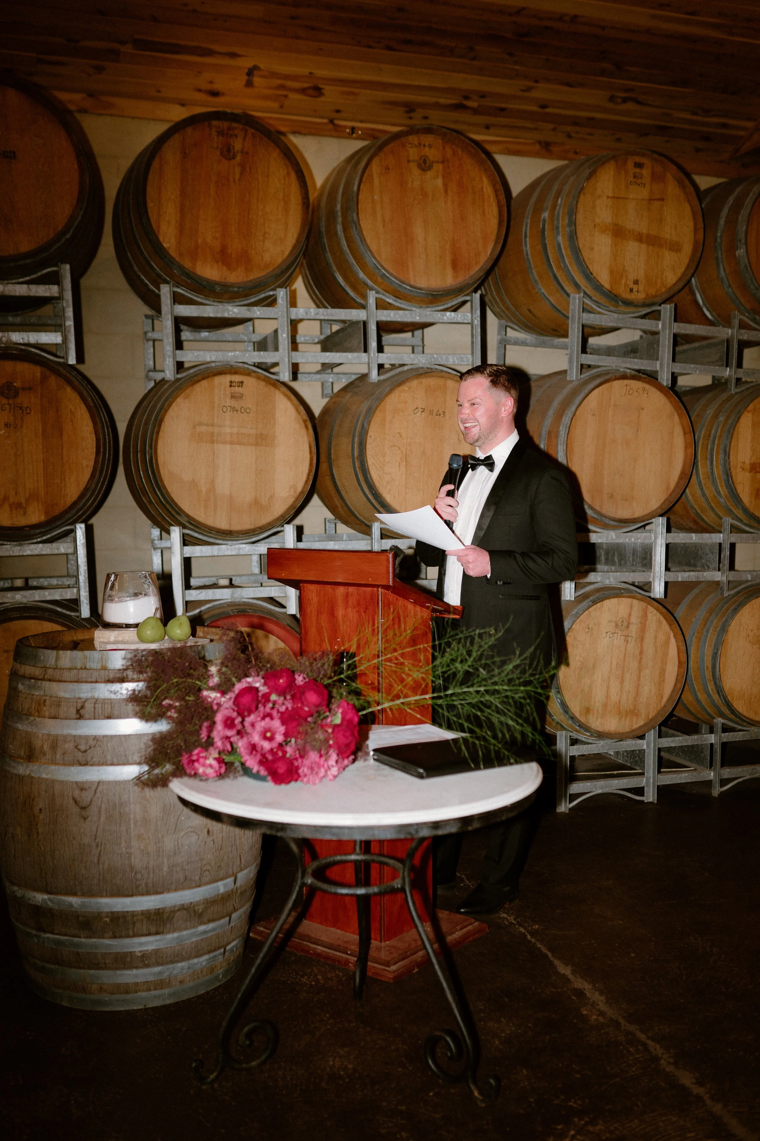 A man in a black tuxedo and bow tie giving a speech at a winery or wine cellar, standing at a wooden podium with a microphone, surrounded by wine barrels, with a floral arrangement and a glass of water on a barrel in front.