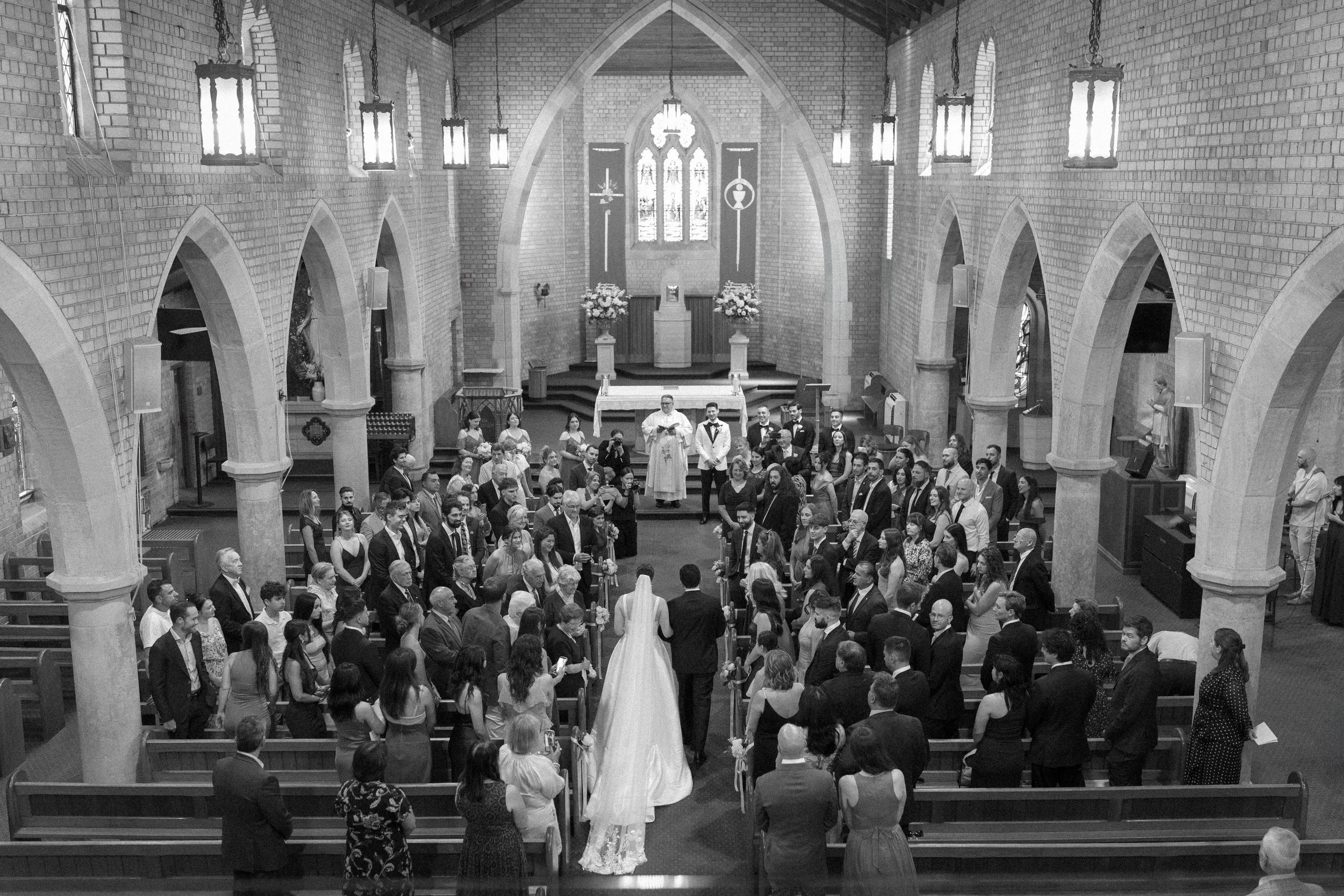 A wedding ceremony inside a church with the bride and groom at the altar. Guests are seated in pews, watching the couple, and a priest is leading the service.