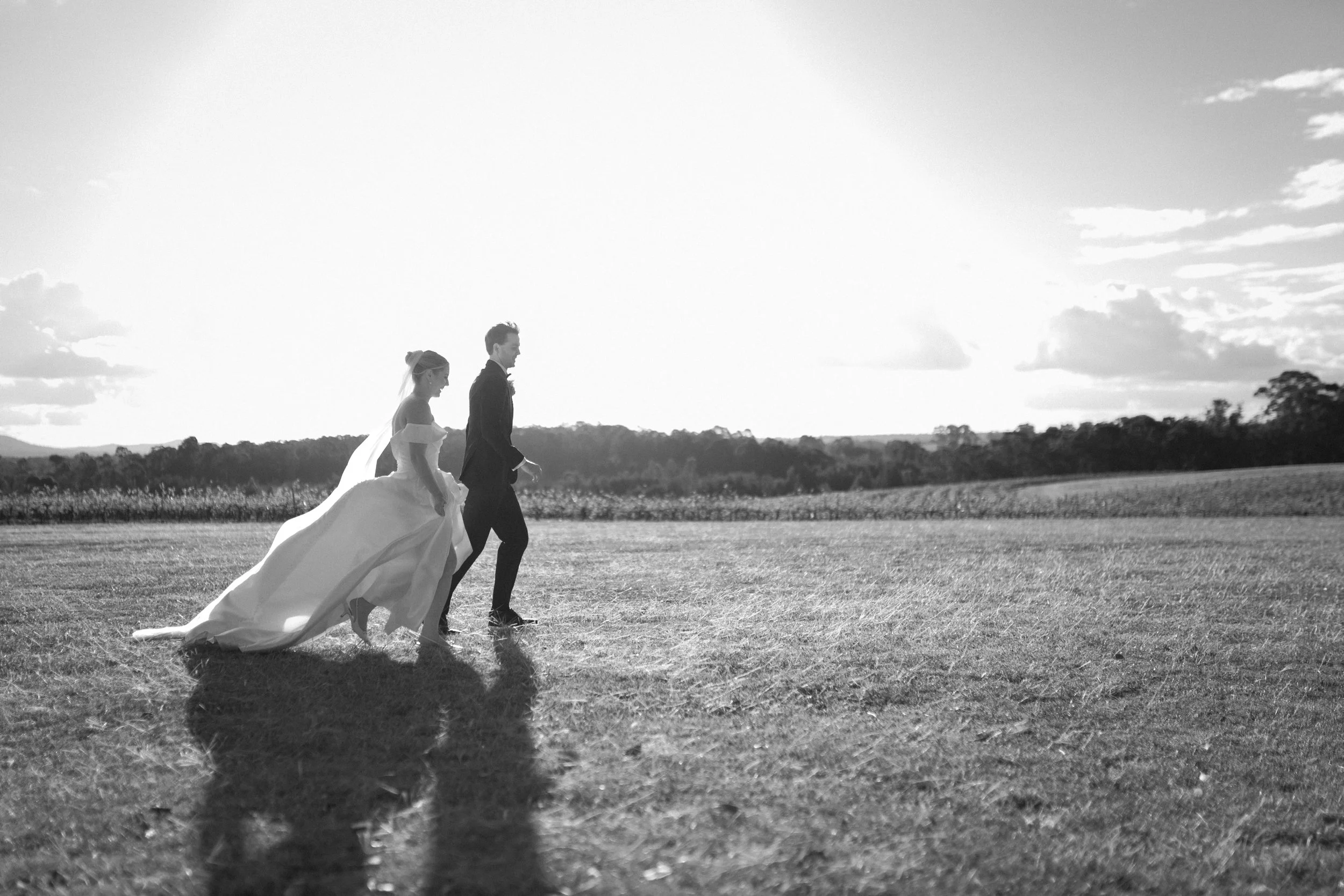 Black and white photo of a bride and groom walking outdoors across an open field, with a scenic background of trees and clouds in the sky. Bimbadgen Palmers Lane Wedding, Hunter Valley