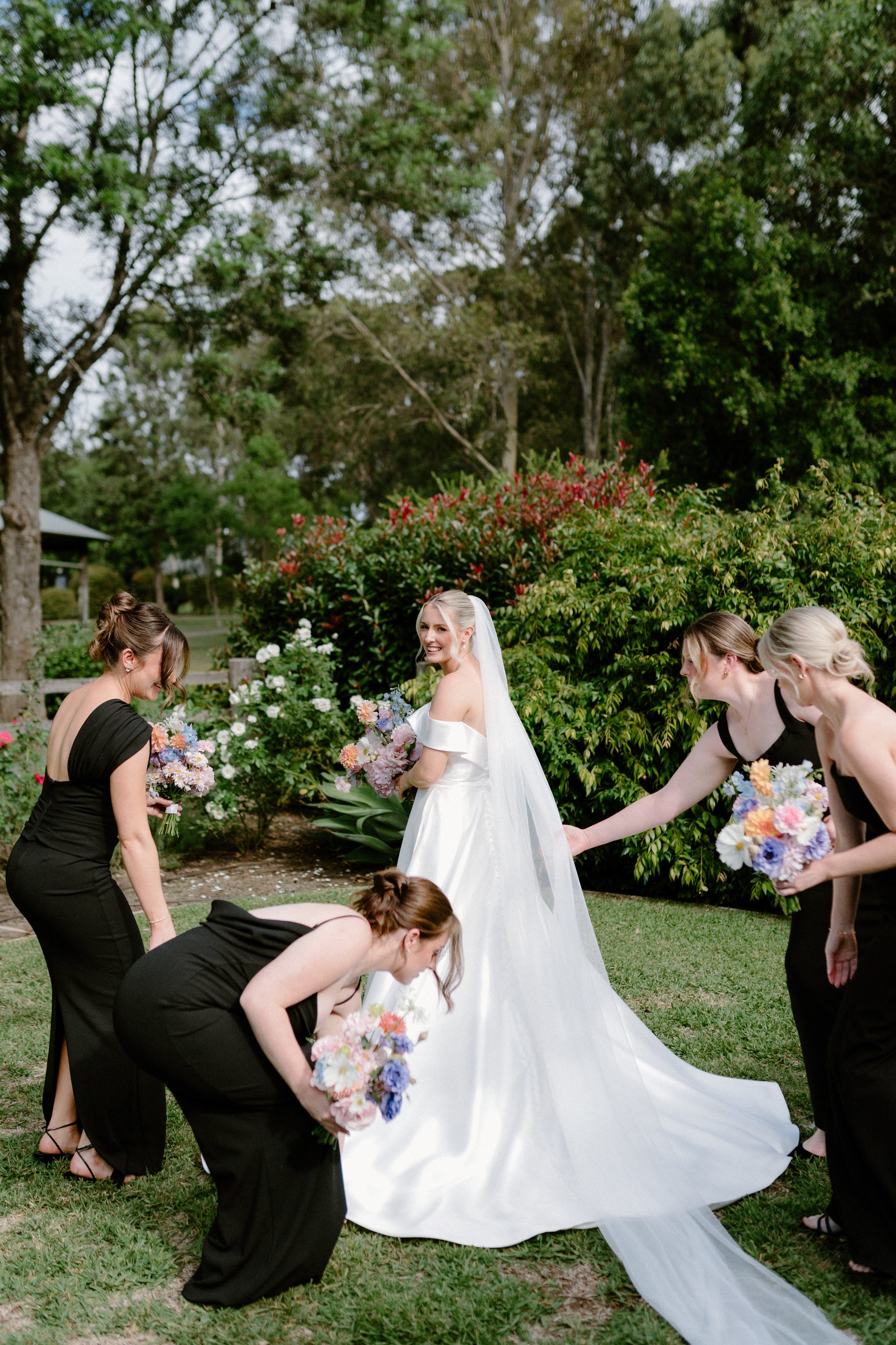 Bride in a white wedding gown and veil holding a bouquet, surrounded by bridesmaids in black dresses with bouquets, outside in a garden with trees and bushes.