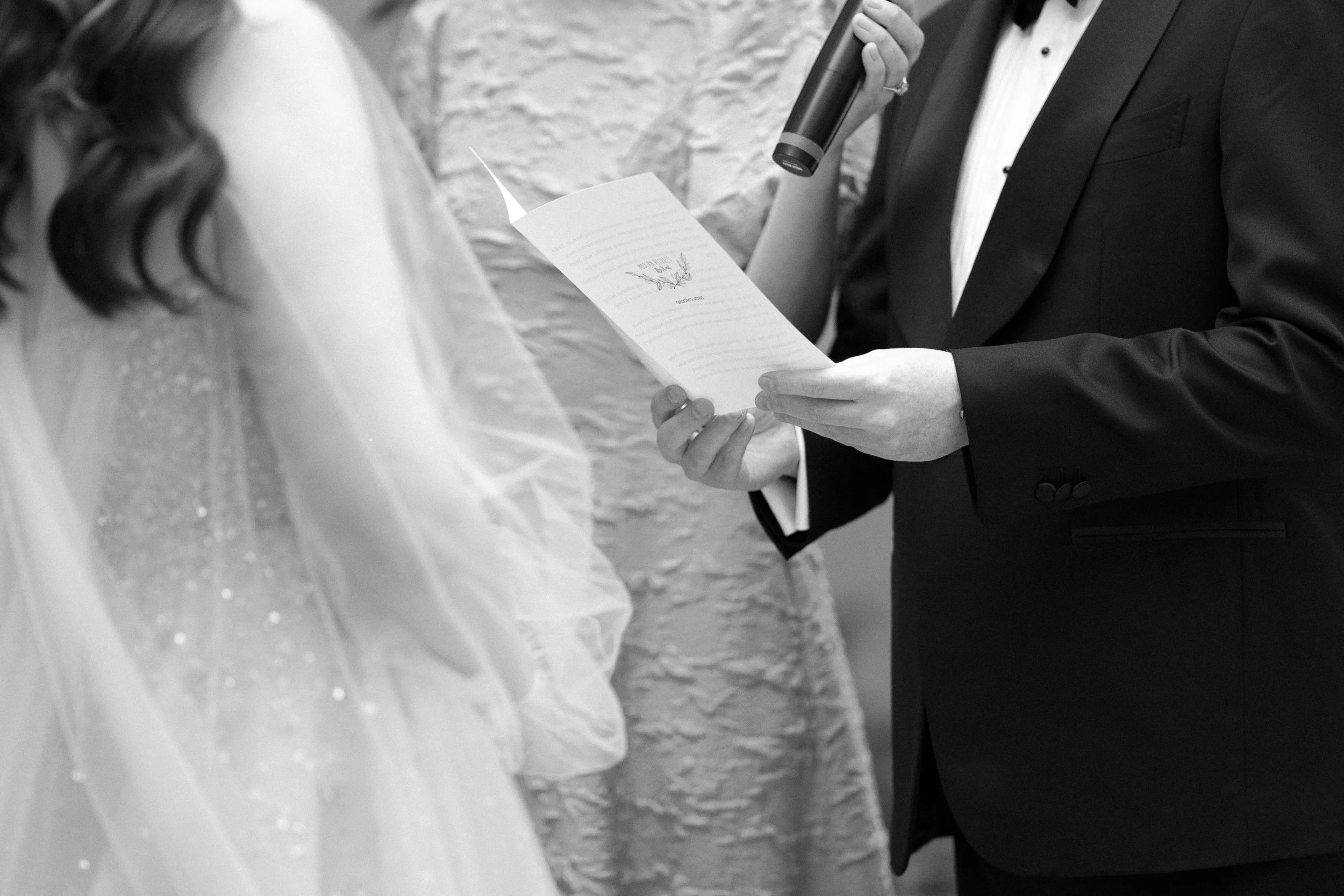A groom in a tuxedo reading vows from a paper during a wedding ceremony.