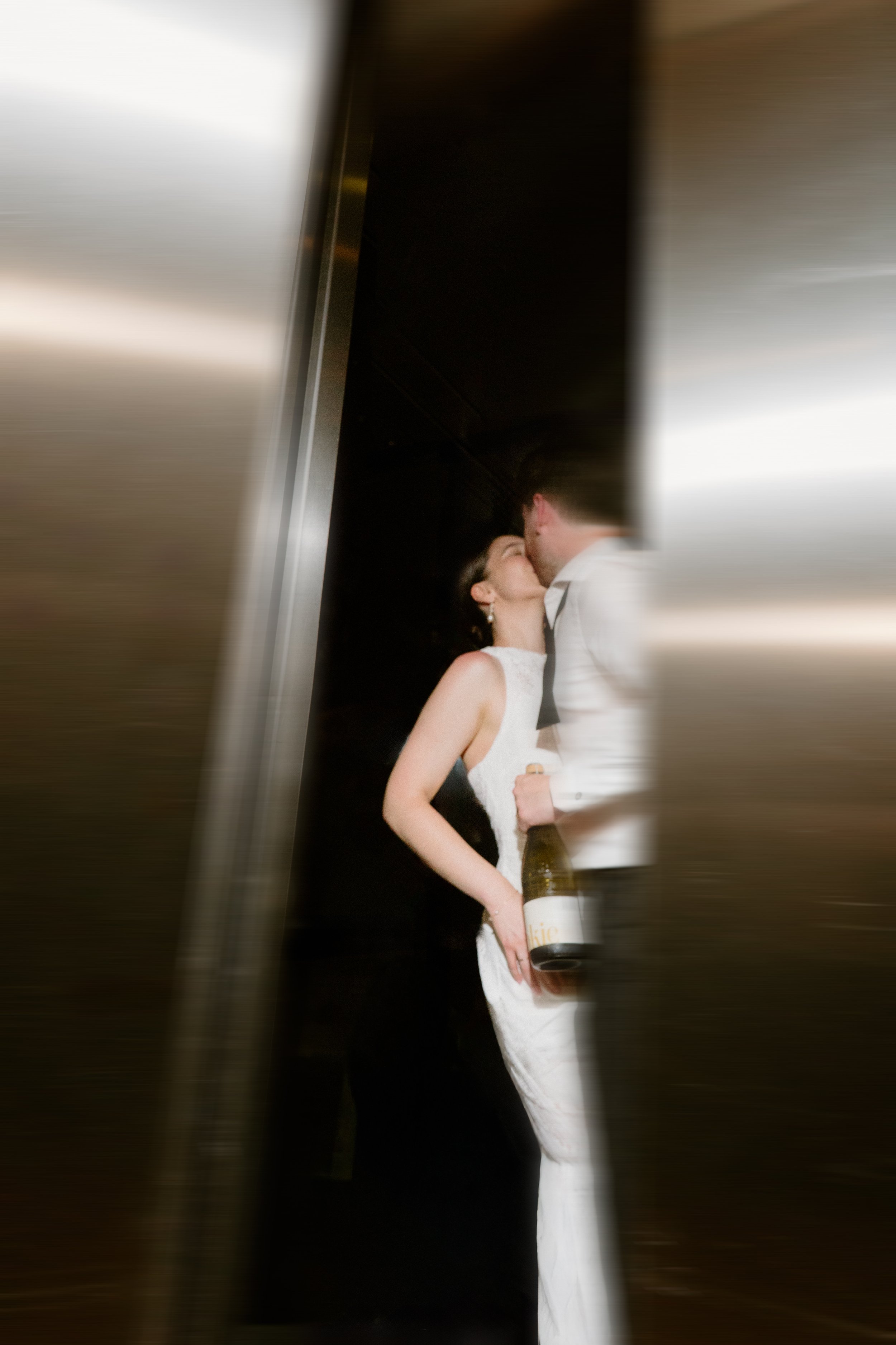 Sergeants Mess, Mosman Wedding Venue. Sergeants Mess Wedding Photos. Couple in formal attire sharing a kiss, with the woman holding a bottle of champagne, seen through a partially open elevator door.