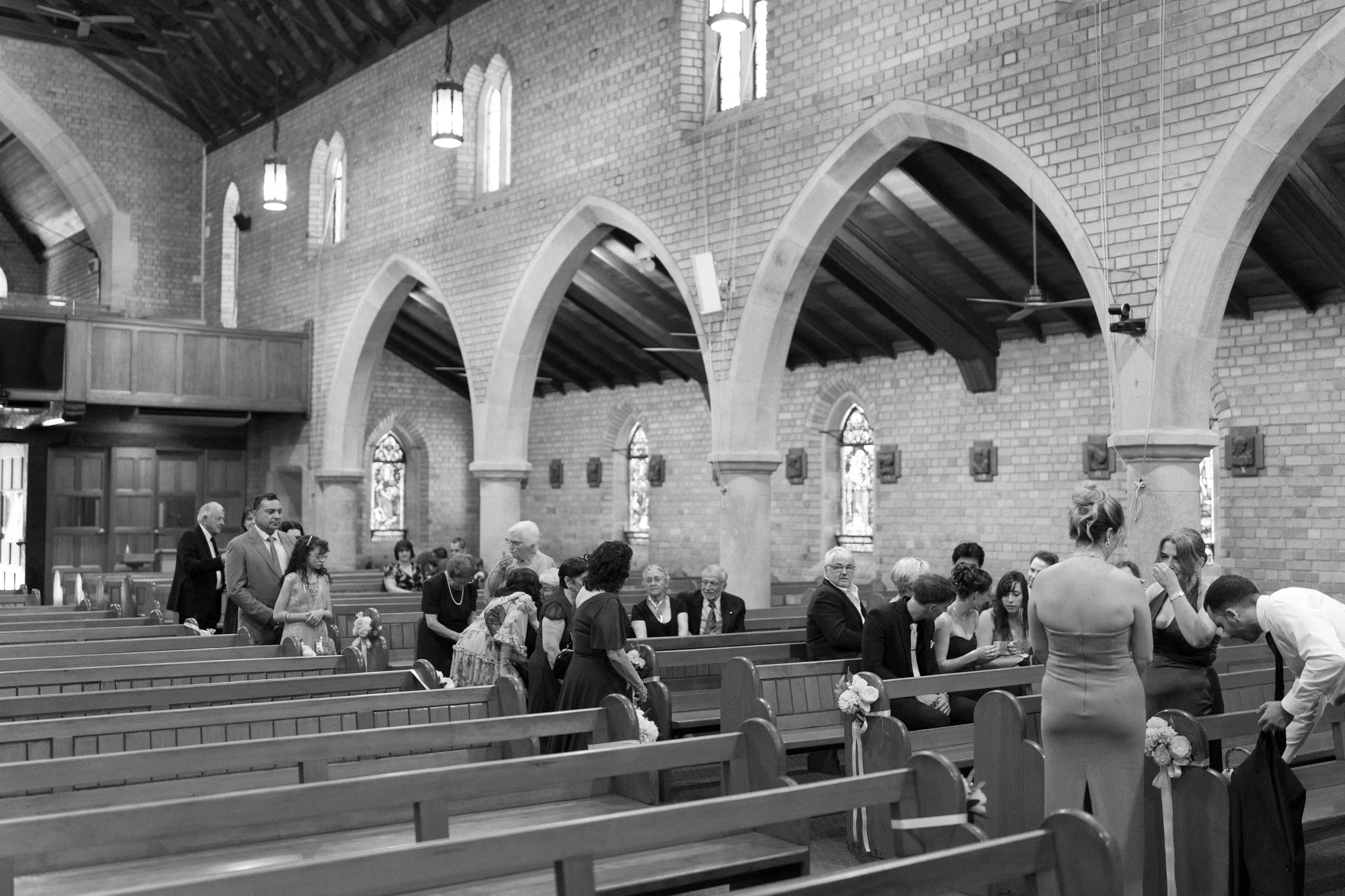 People gathered inside a church with brick walls and stained glass windows, some sitting on pews and others standing or talking, likely before a wedding or ceremony.