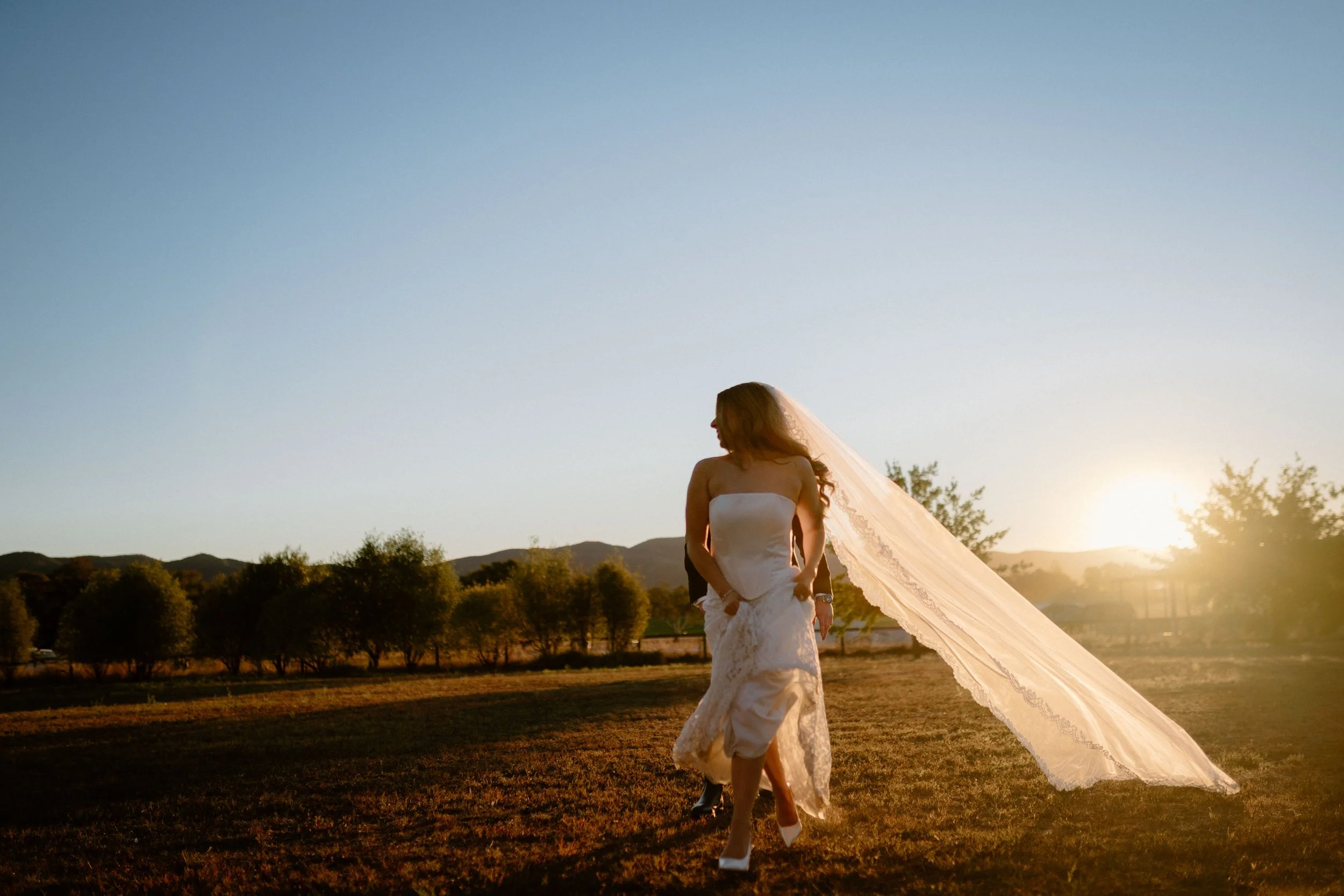 A bride in a white strapless wedding dress and veil walking outdoors in a field during sunset.