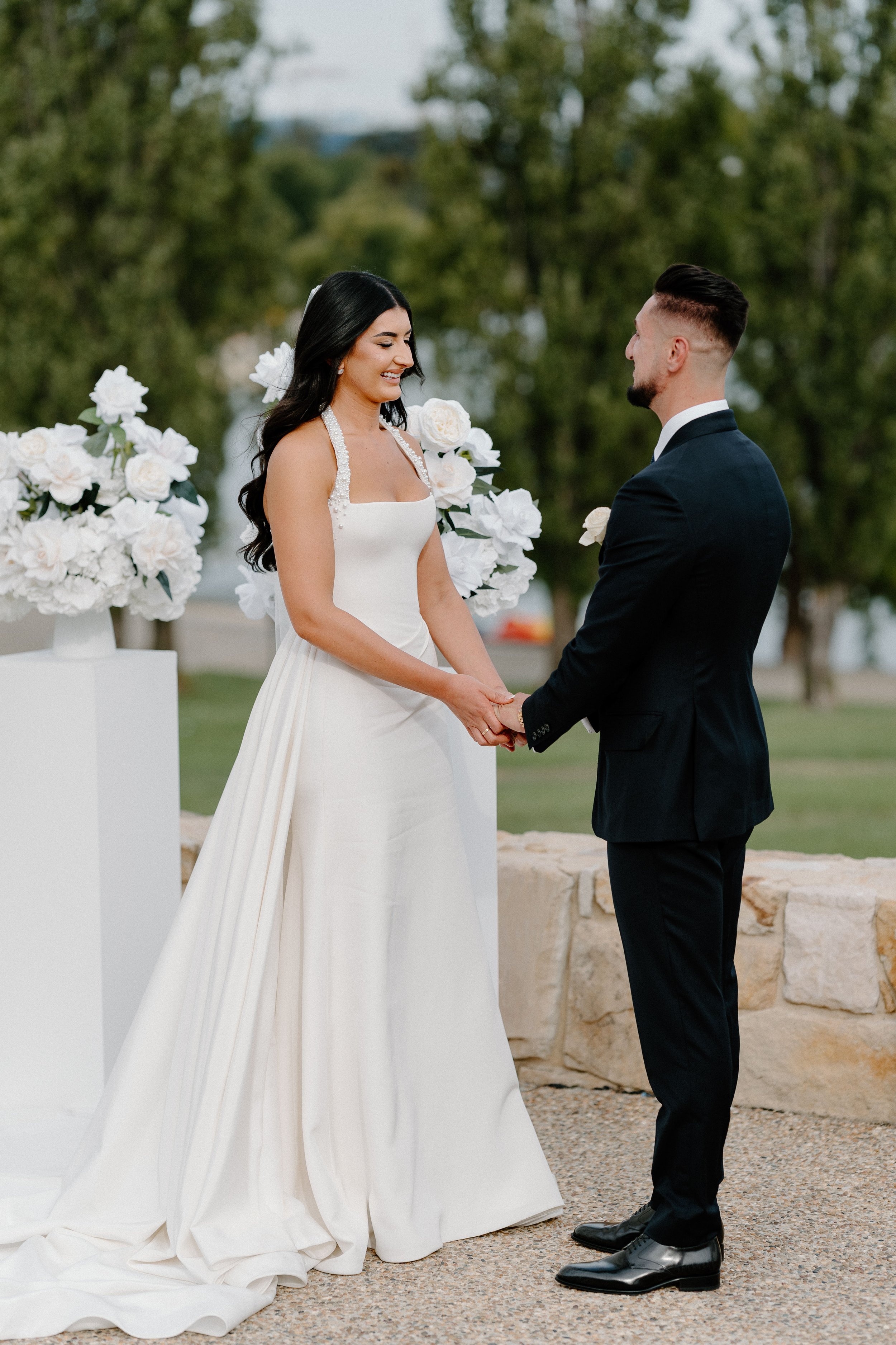 A bride and groom holding hands and smiling during their outdoor wedding ceremony with white floral arrangements in the background.