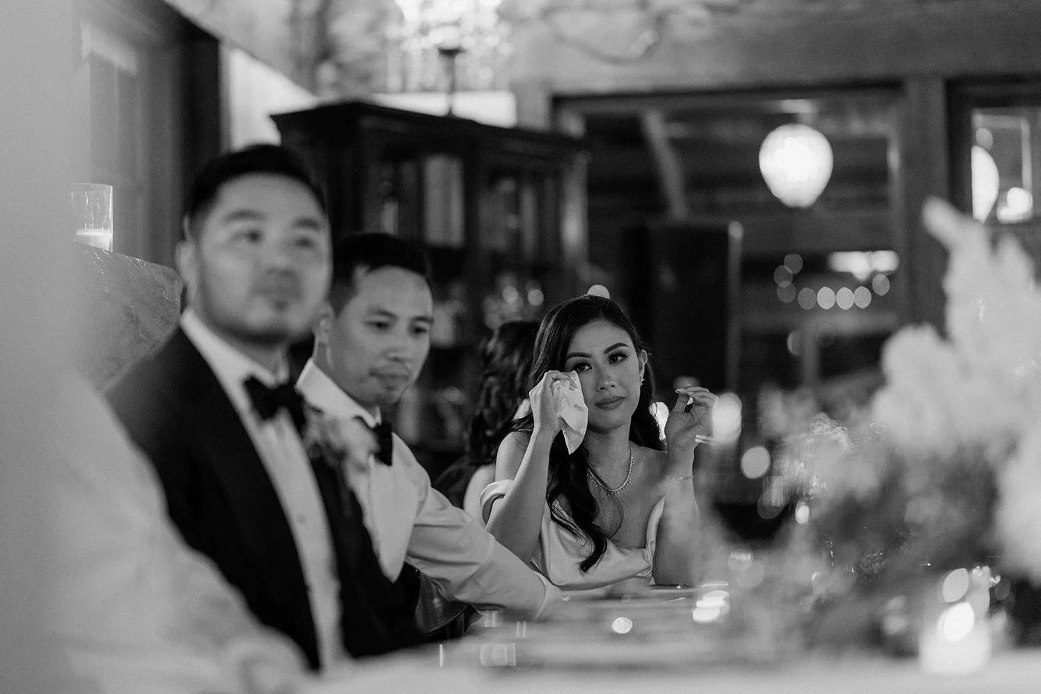 Black and white photo of a formal event with people sitting at a table, including a woman wiping her eye with a tissue, surrounded by men in tuxedos.