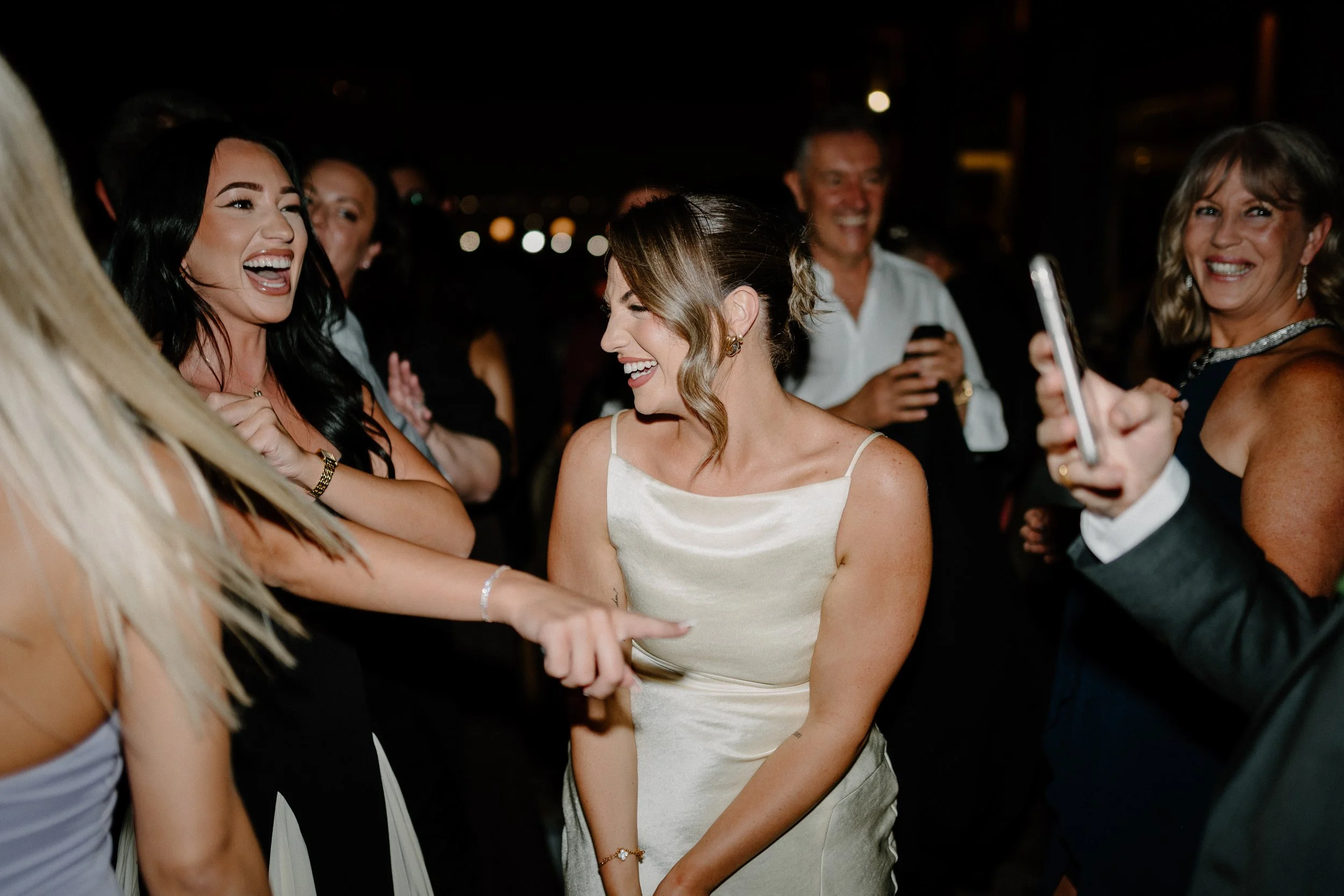 Group of people smiling and laughing at a celebration, including a woman in a white dress with brown hair styled in loose curls.