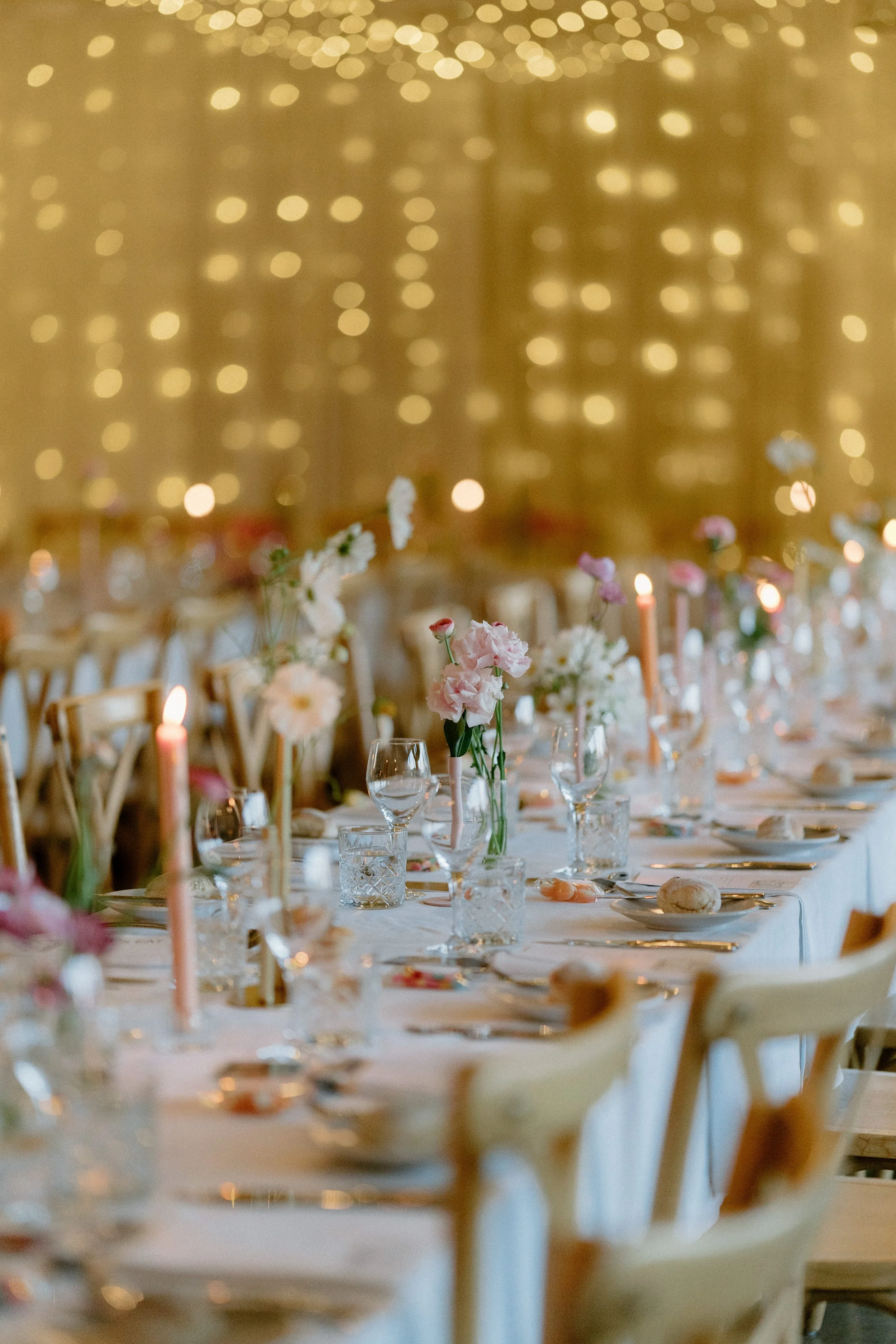 Elegant table setting with flowers, candles, and glassware, decorated for a celebration or wedding reception, against a backdrop of warm, golden fairy lights.