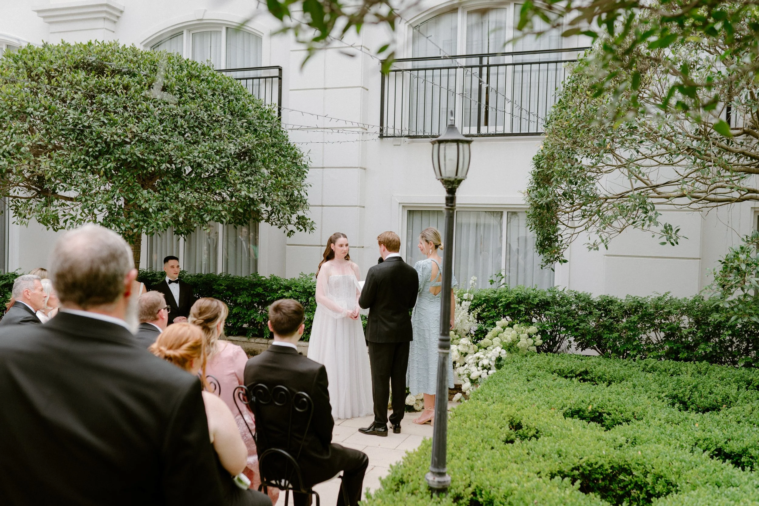 A wedding ceremony taking place outdoors in a garden courtyard. The bride and groom are standing facing each other, with an officiant standing nearby. Guests are seated, watching the ceremony, with some dressed in formal attire. The setting includes 