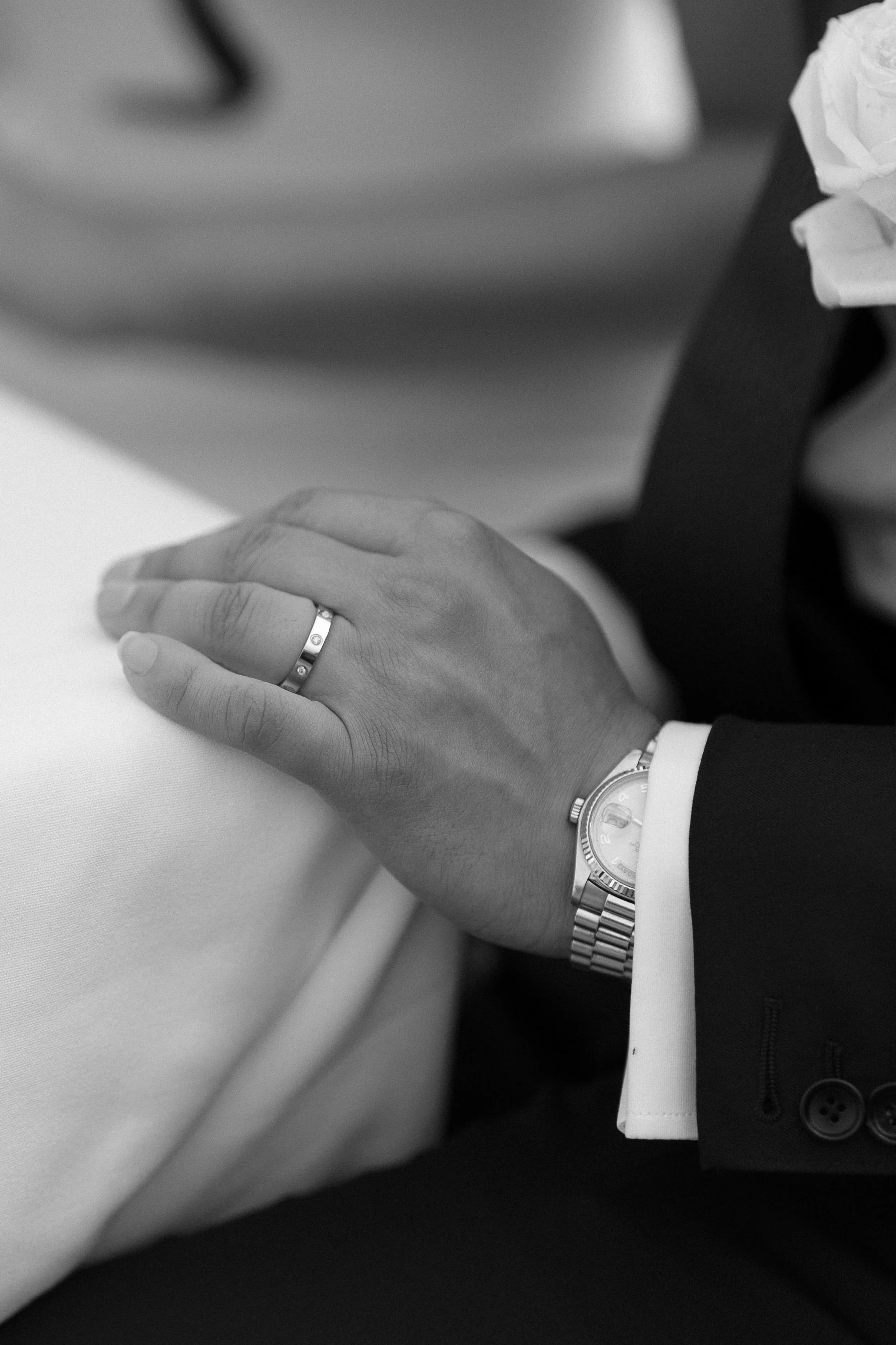 Close-up of a person wearing a wedding ring and a wristwatch, with a white shirt cuff visible, dressed in a dark suit. Someone is sitting at a table with a white tablecloth.